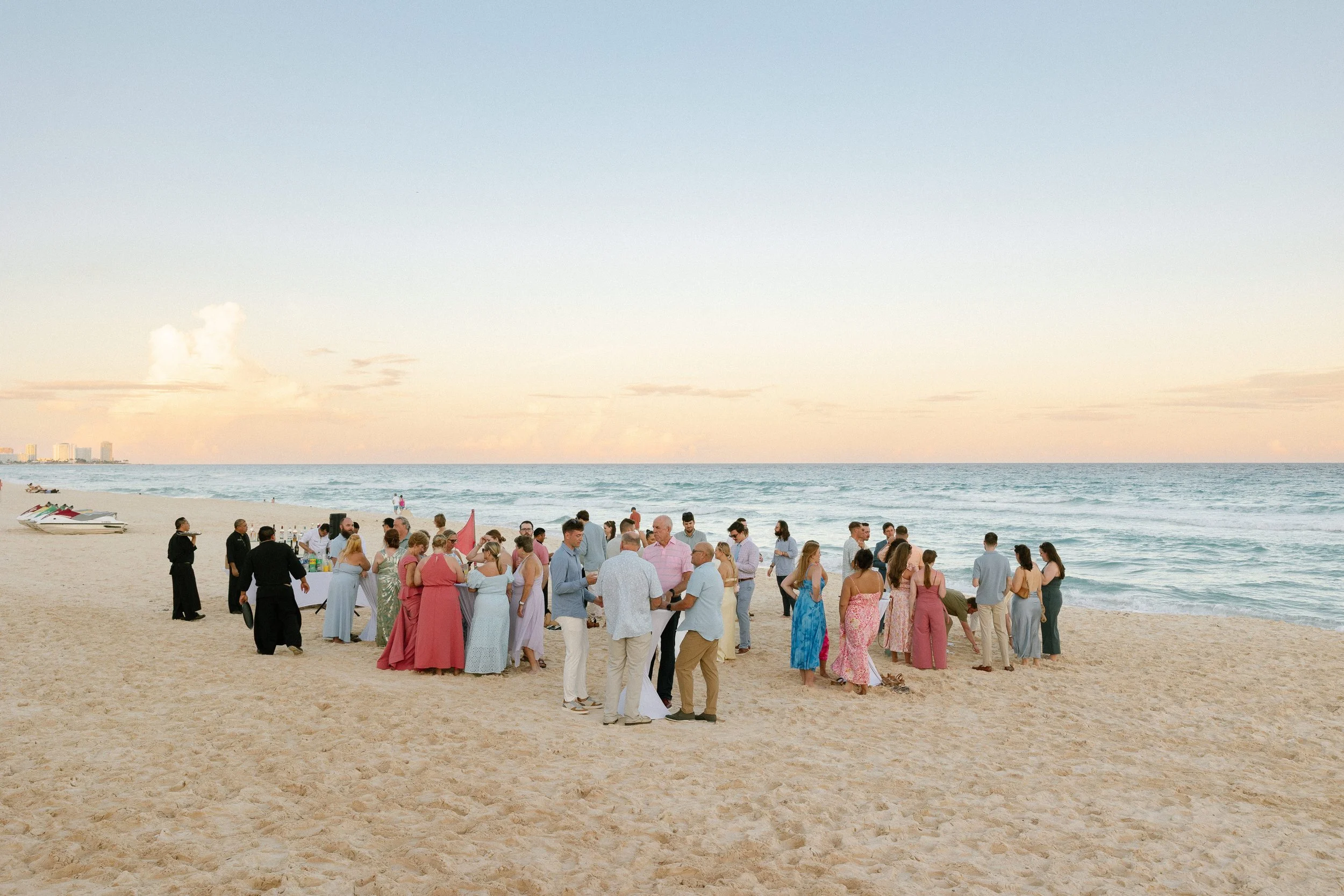 A large group of people dressed in fancy clothes gathering on a sandy beach for a celebration or event, with ocean waves and a pastel sky in the background.
