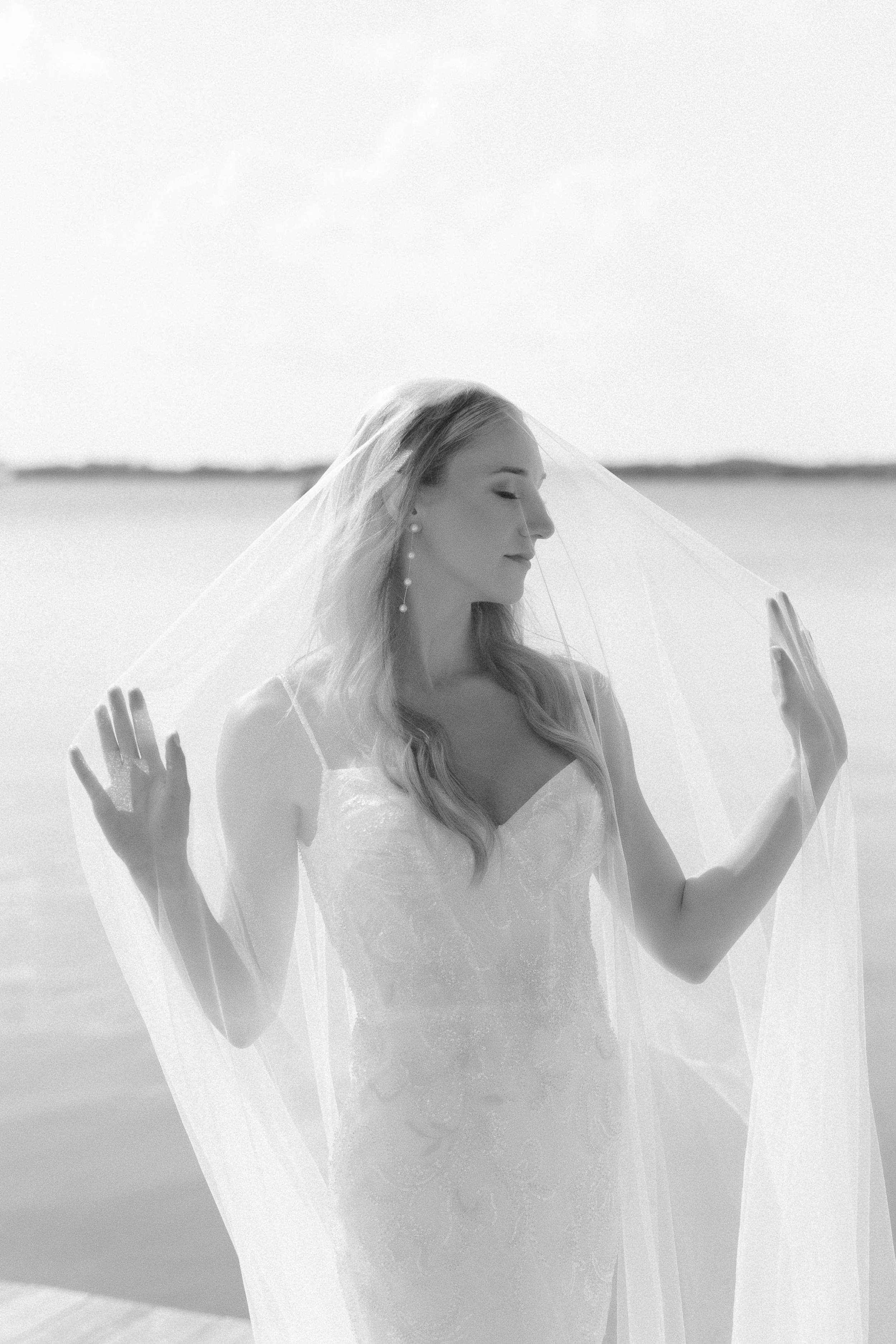 A woman with long hair and pearl earrings, with a veil over her face, standing outdoors in front of water with a calm, serene expression, her eyes closed and hands gently raised.