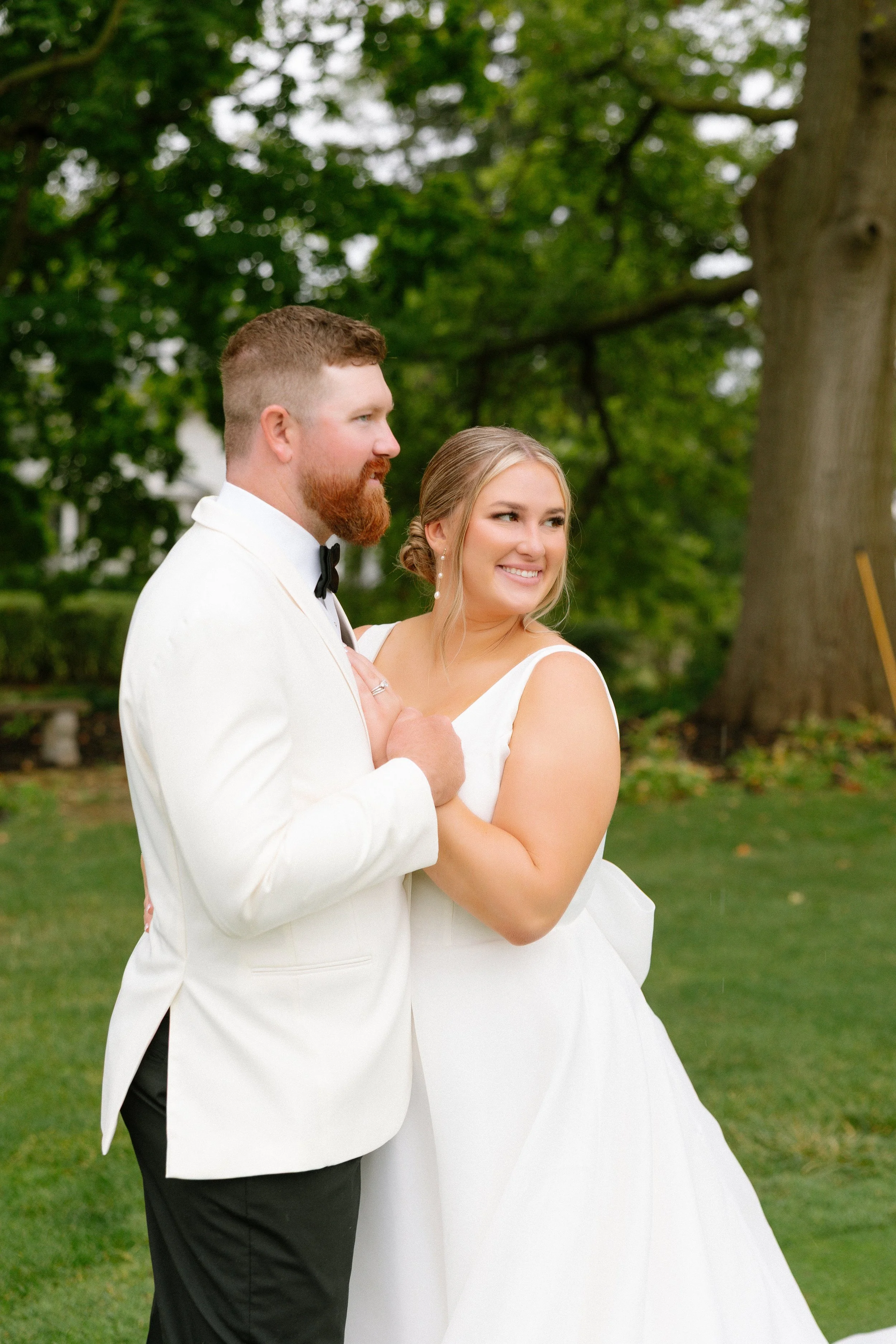 A bride and groom standing outdoors on a grassy area with trees in the background, smiling and holding each other.