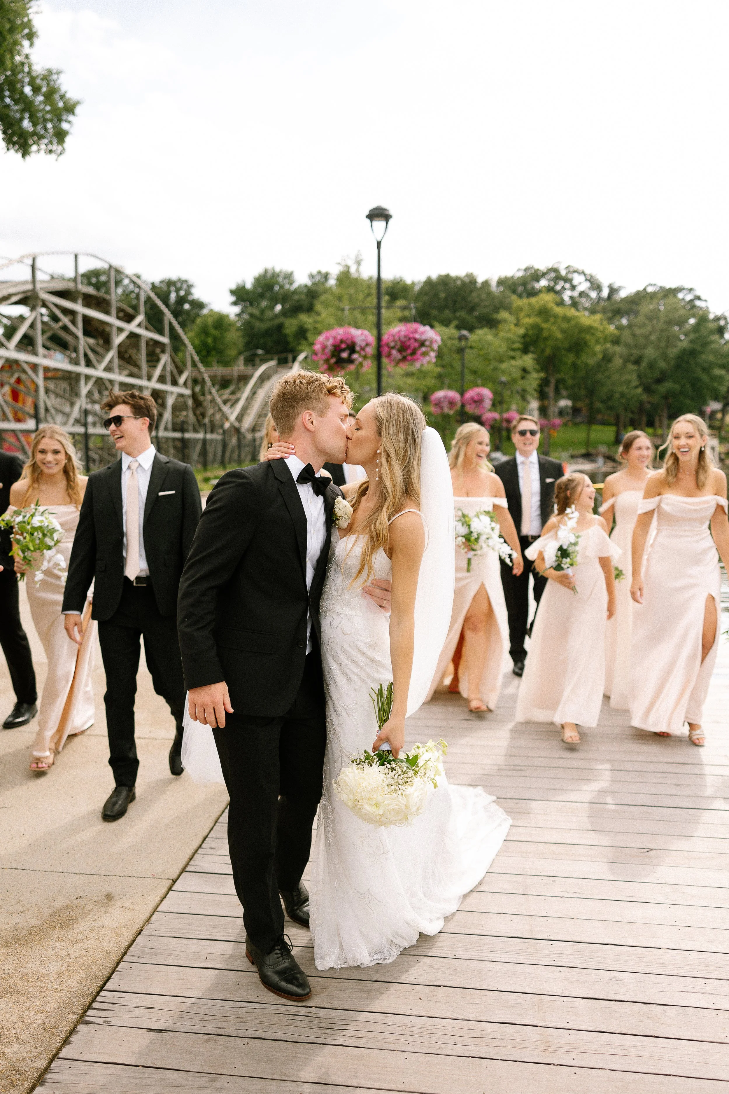 A newlywed couple kisses during their wedding ceremony, with bridesmaids and groomsmen smiling in the background at an outdoor venue decorated with pink flowers and trees.