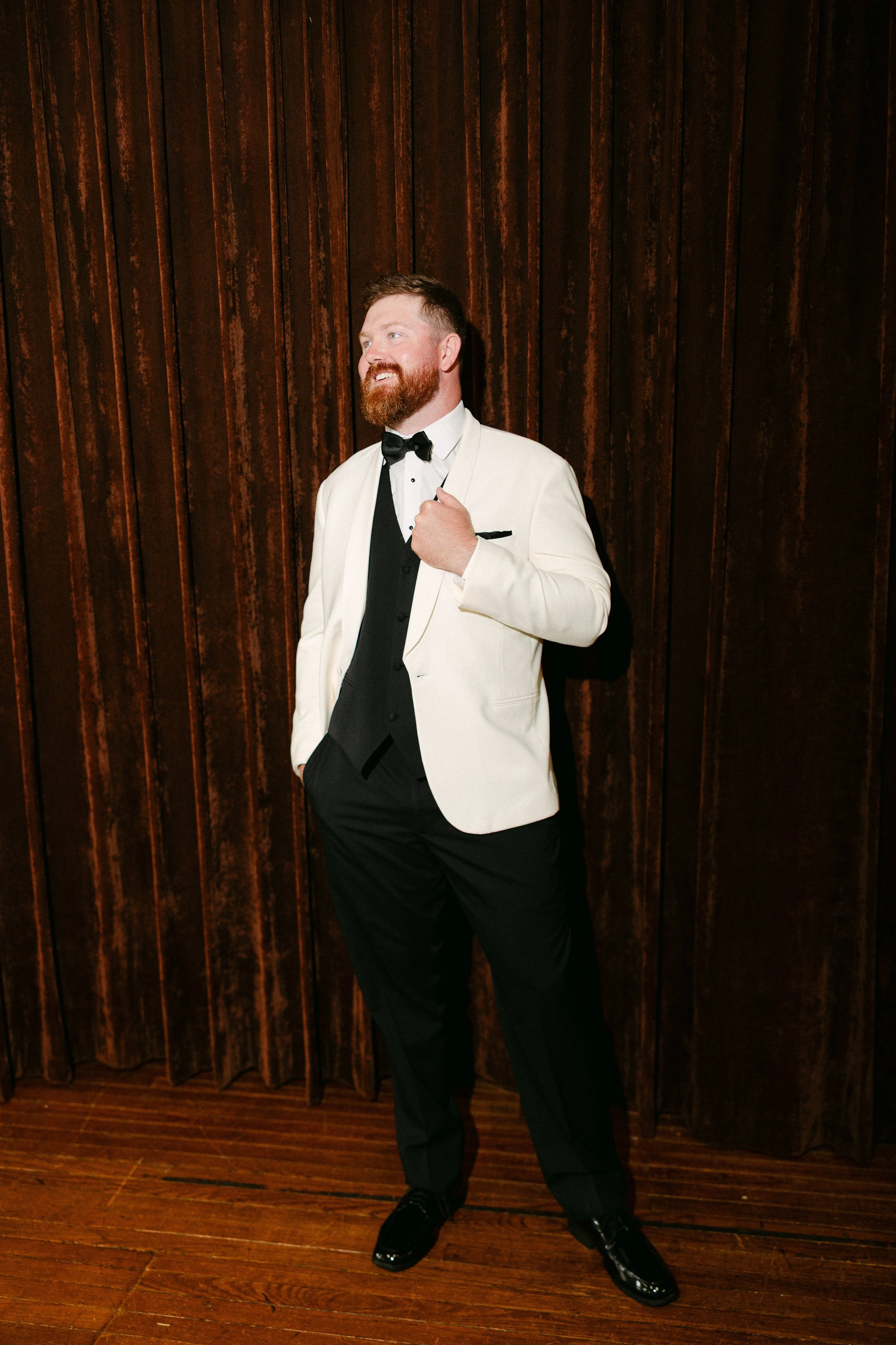A man wearing a white tuxedo jacket, black dress pants, and a black bow tie, standing with one hand in his pocket and the other adjusting his jacket, against a dark wood-paneled background.