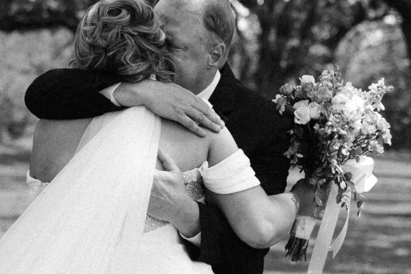 A woman and a man embrace in a hug, with the woman holding a bouquet of flowers. The image is in black and white.