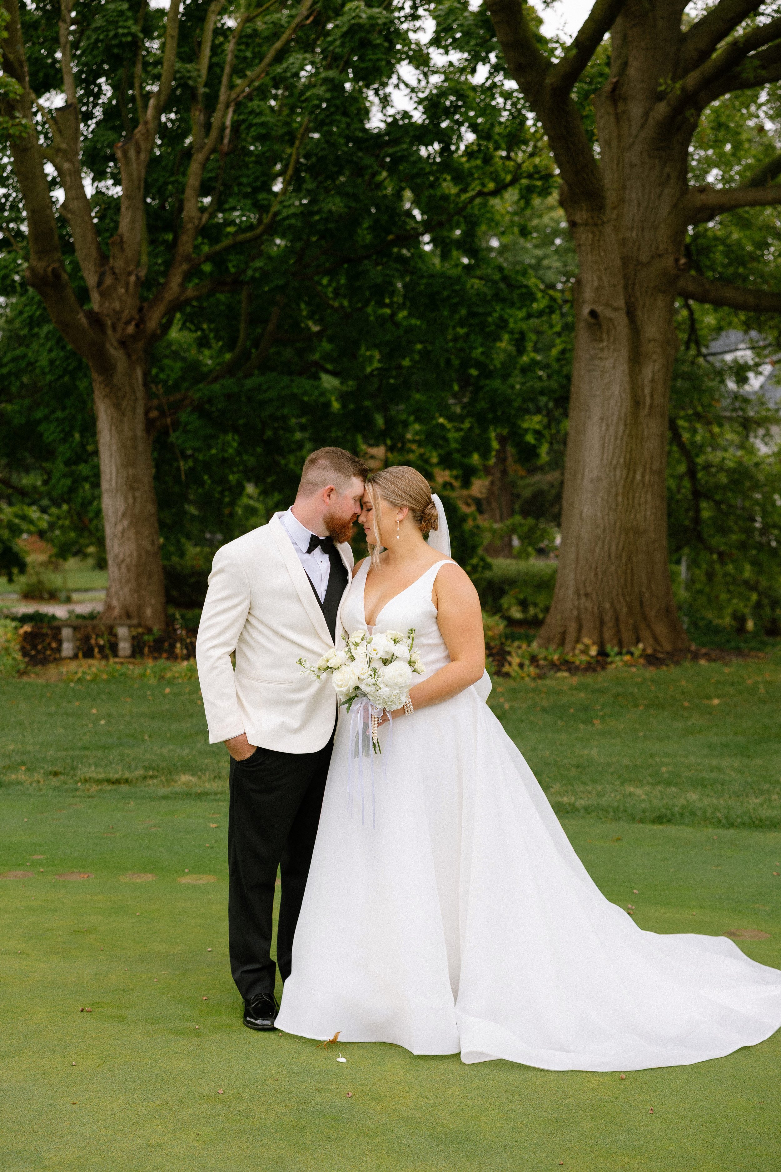 Bride in a white wedding gown and groom in a white tuxedo jacket with black pants, standing close with foreheads touching in a park with large trees.