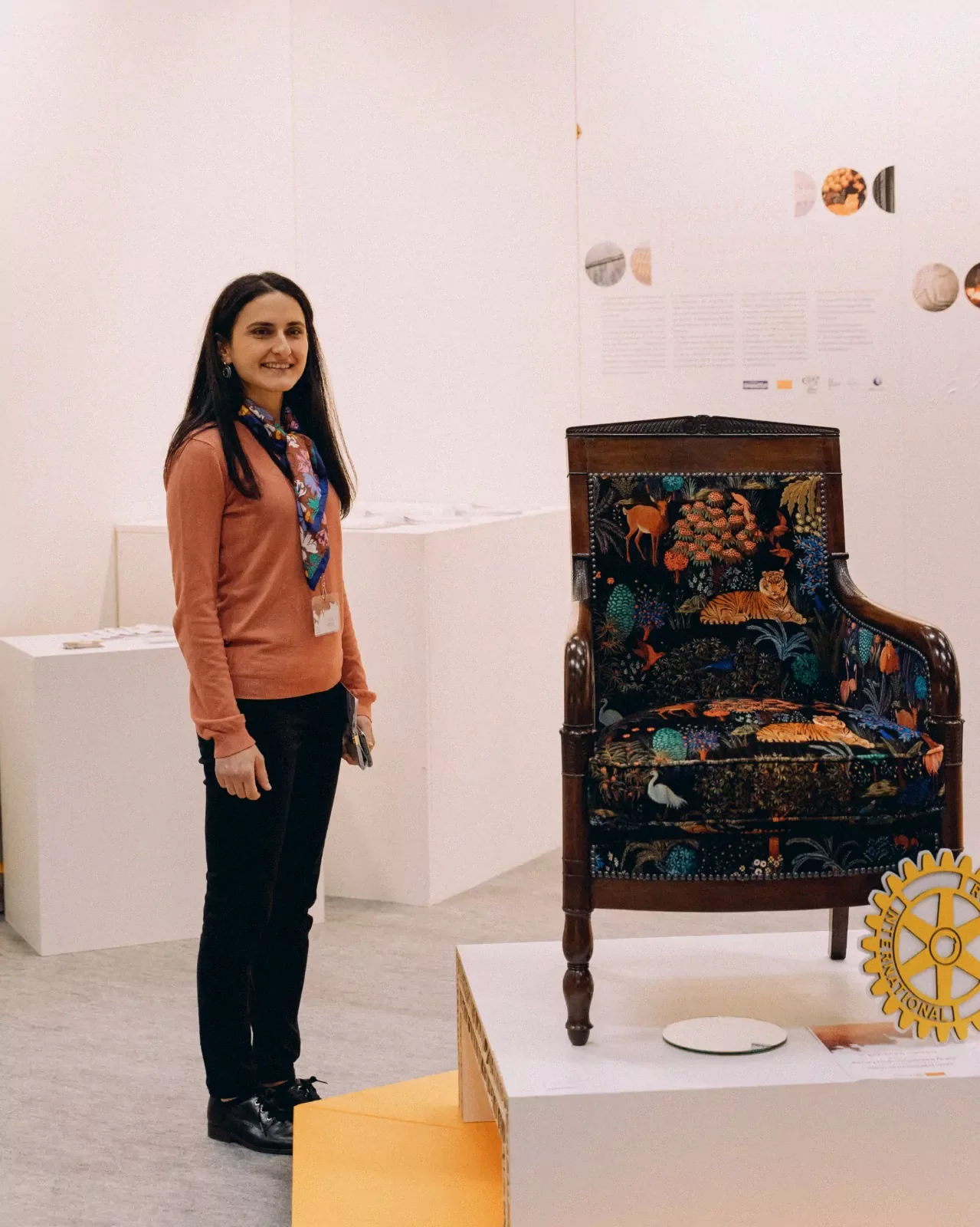 Une femme souriante posant à côté d'un fauteuil en tissu imprimé contenant des animaux sauvages, dans une galerie d'exposition.
