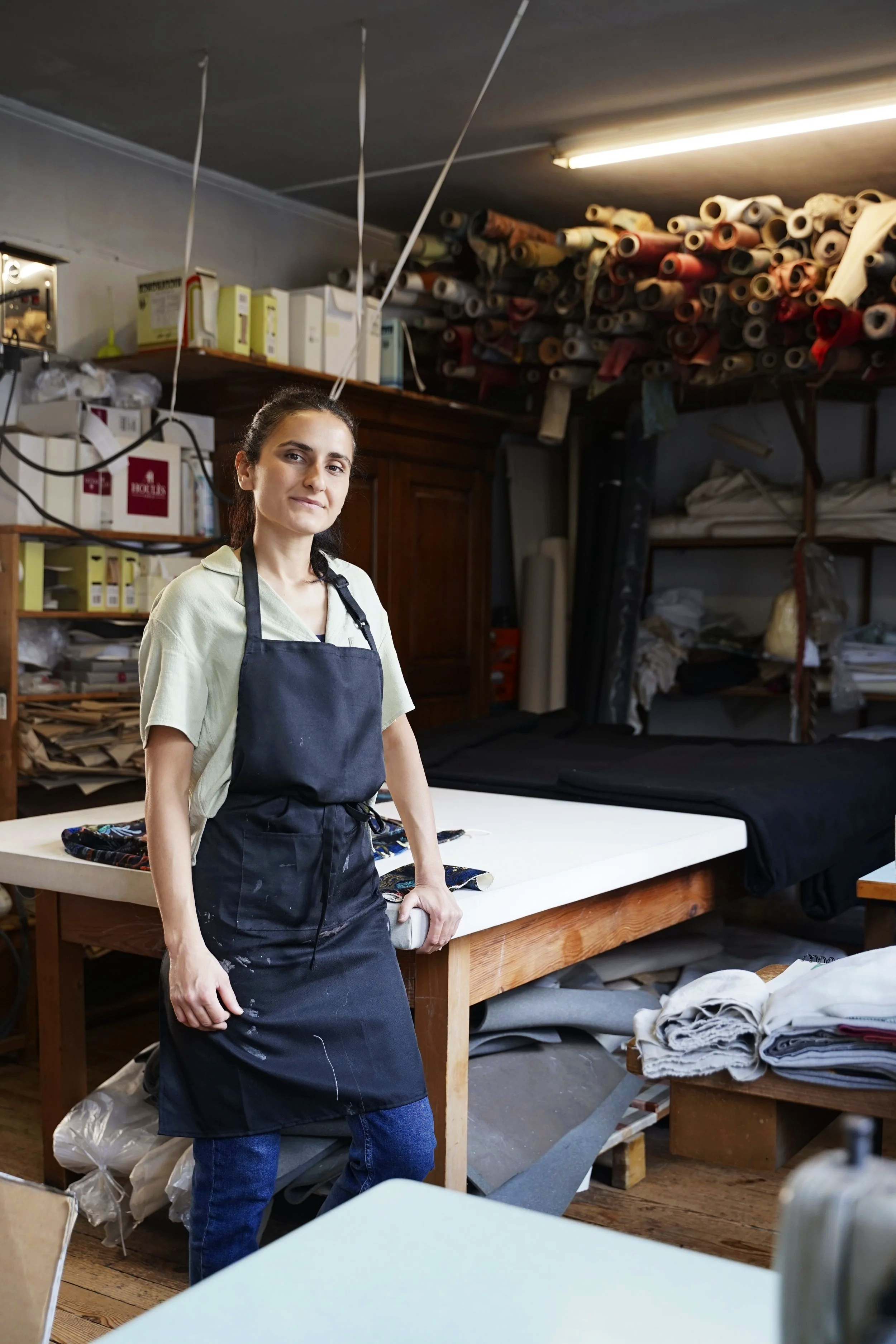 Une femme debout dans un atelier avec des matériaux de couture et des rouleaux de tissus en arrière-plan.