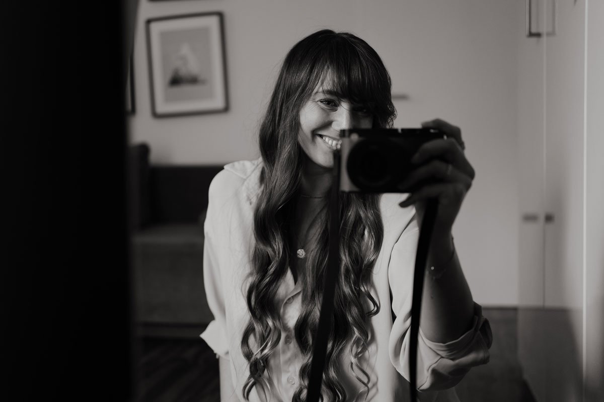 Lächelnde junge Frau mit langen, gewellten Haaren macht ein Selfie mit einer Kamera.