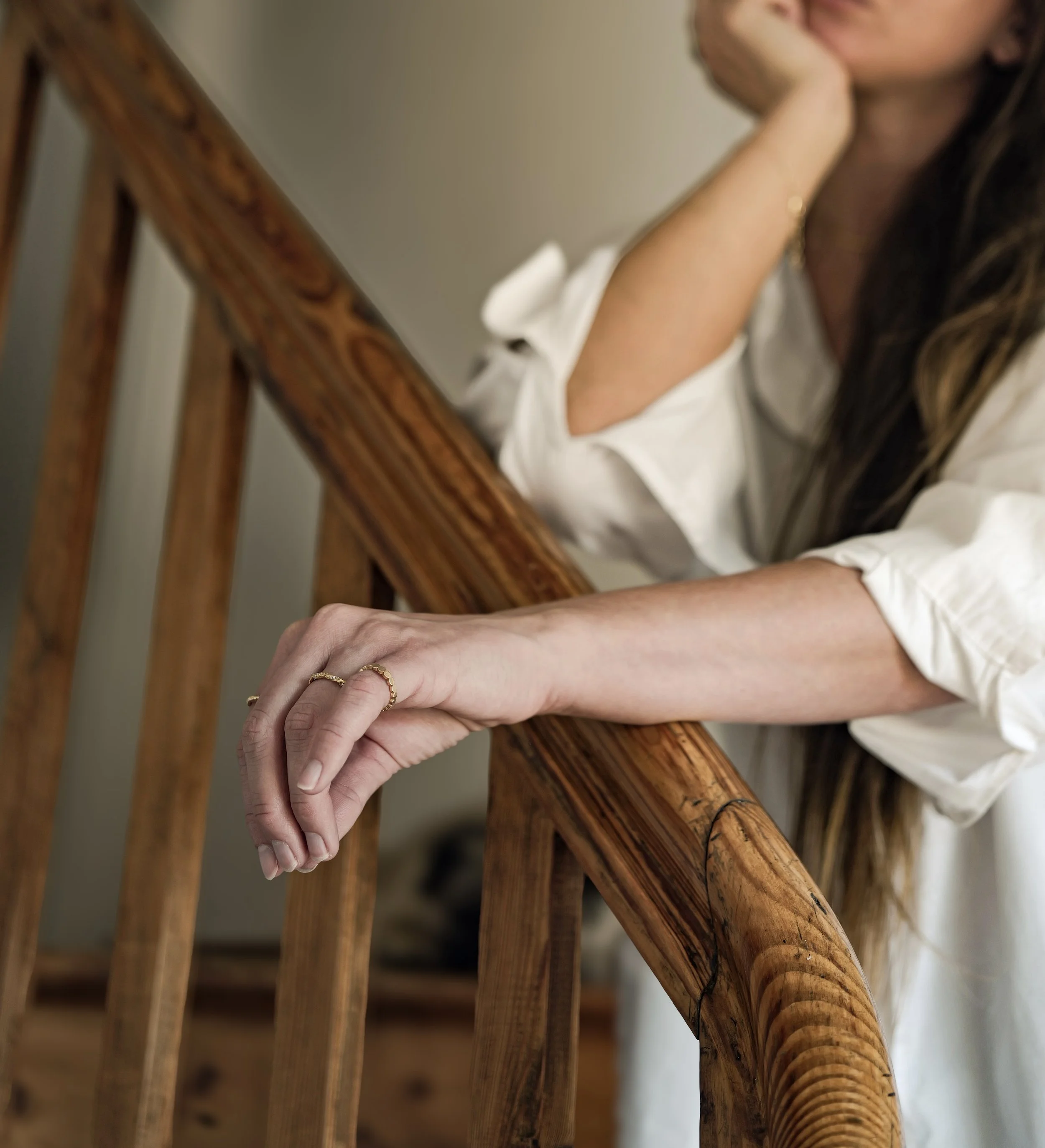 Nahaufnahme einer Hand, die an einem Holzgeländer lehnt, während eine Person im Hintergrund auf einer Treppe sitzt, mit einem weißen Hemd. Die Person trägt einen goldenen Ring und hat lange braune Haare.