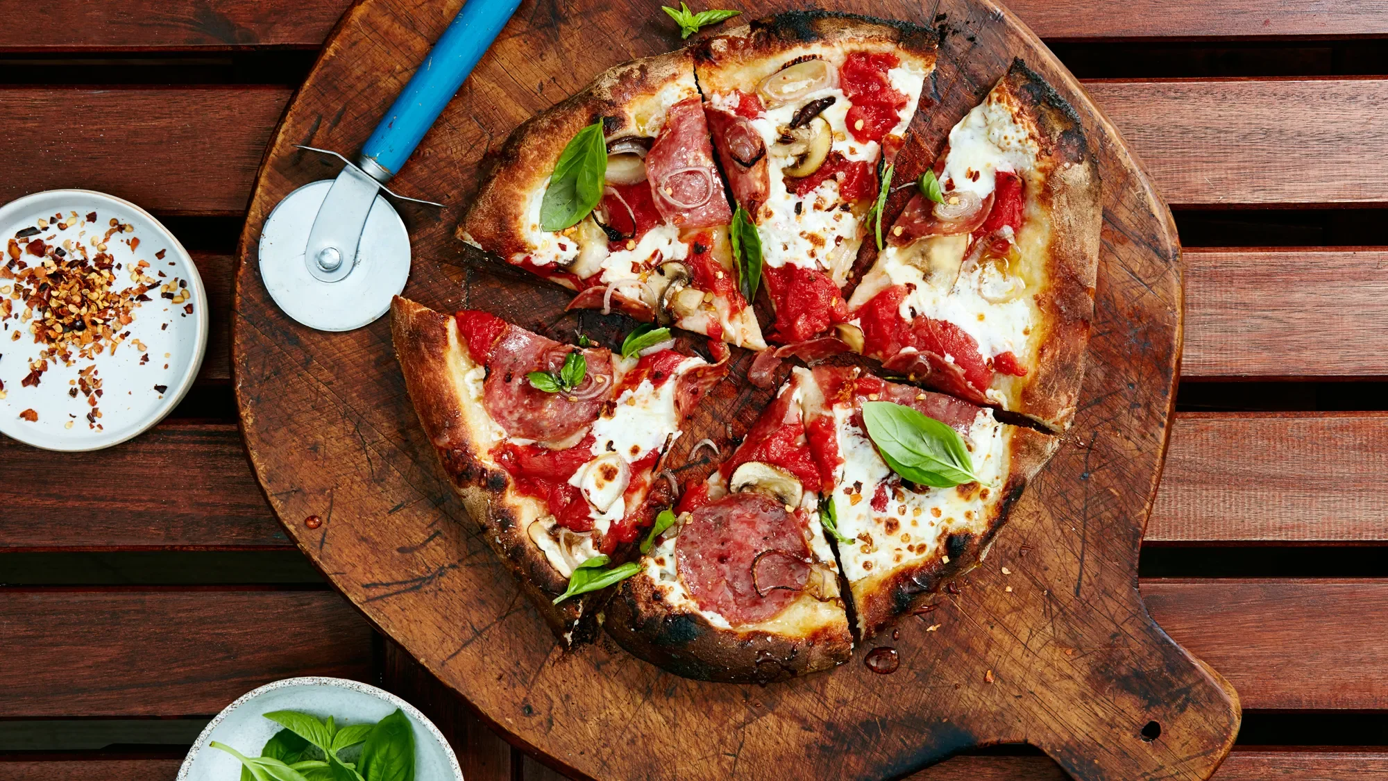 A freshly baked pizza with pepperoni, mushrooms, tomatoes, and basil leaves on a round wooden serving board, with a pizza cutter and small bowls of chopped chili flakes and basil on a wooden table.