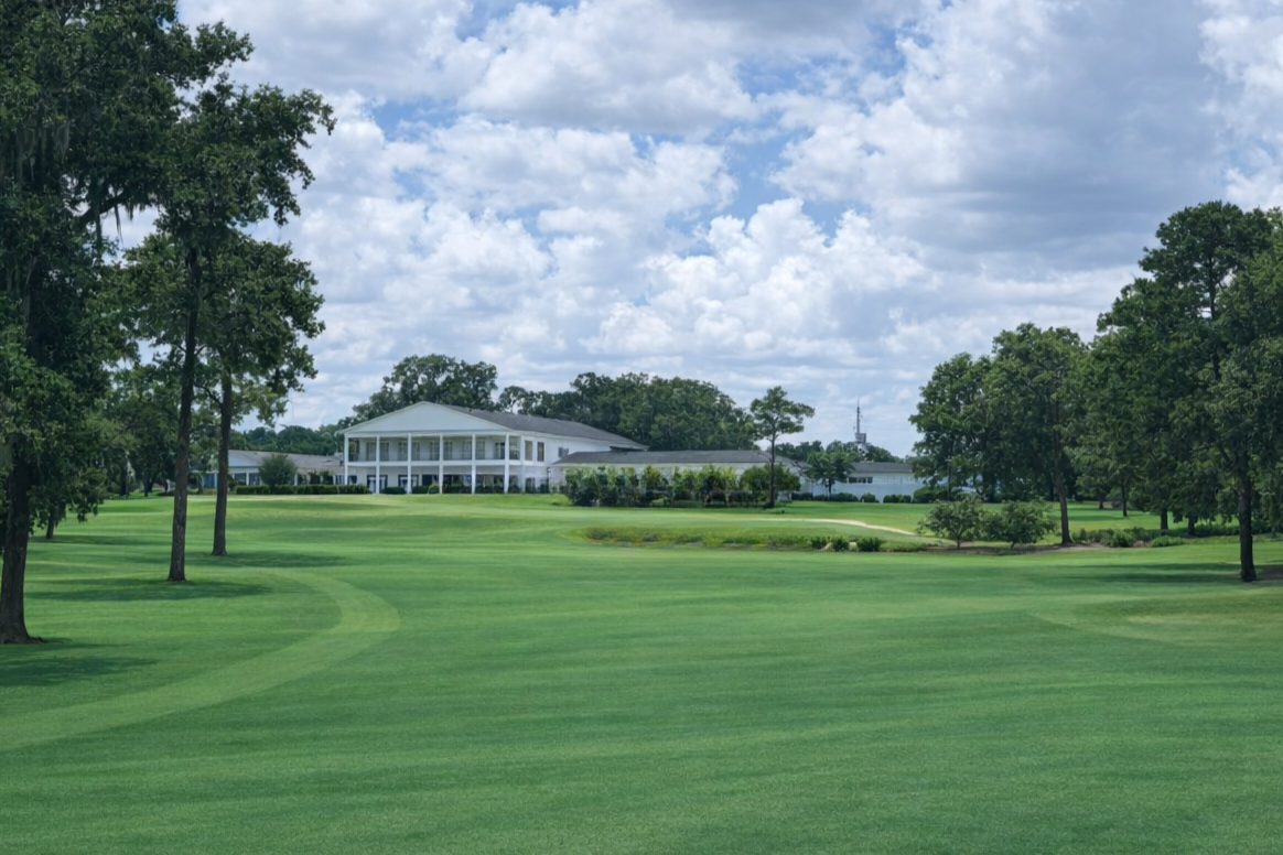 A scenic golf course with well-maintained green grass, scattered trees, and a large white clubhouse building in the background under partly cloudy skies.