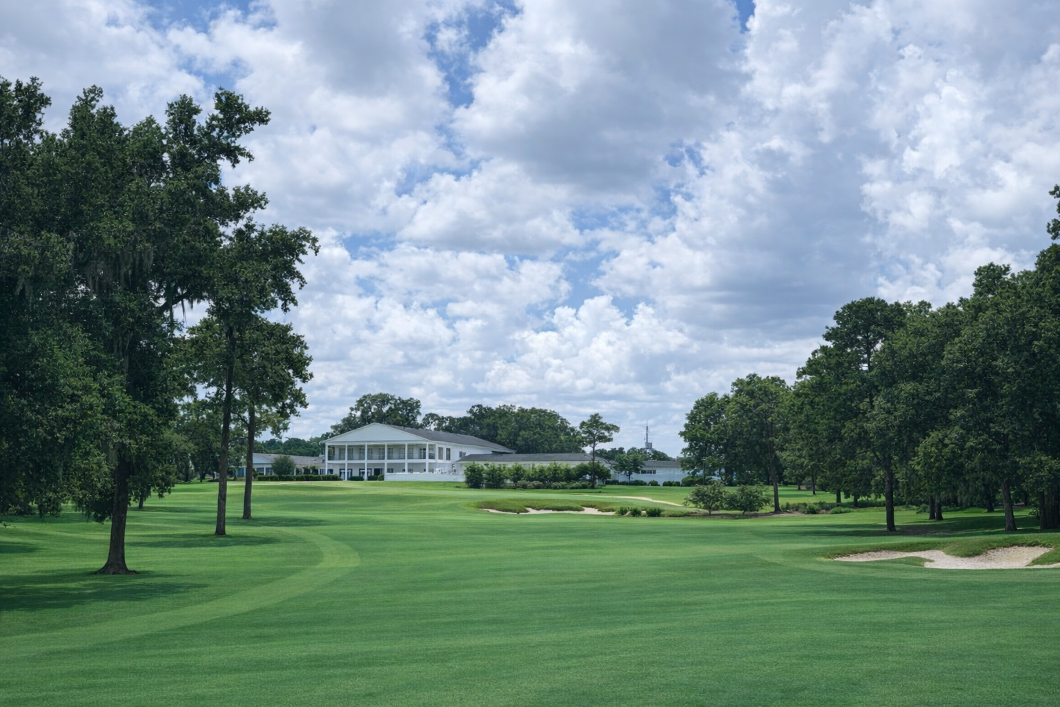 A golf course with lush green grass, sand bunkers, and tall trees, with a large white clubhouse and a partly cloudy sky in the background.