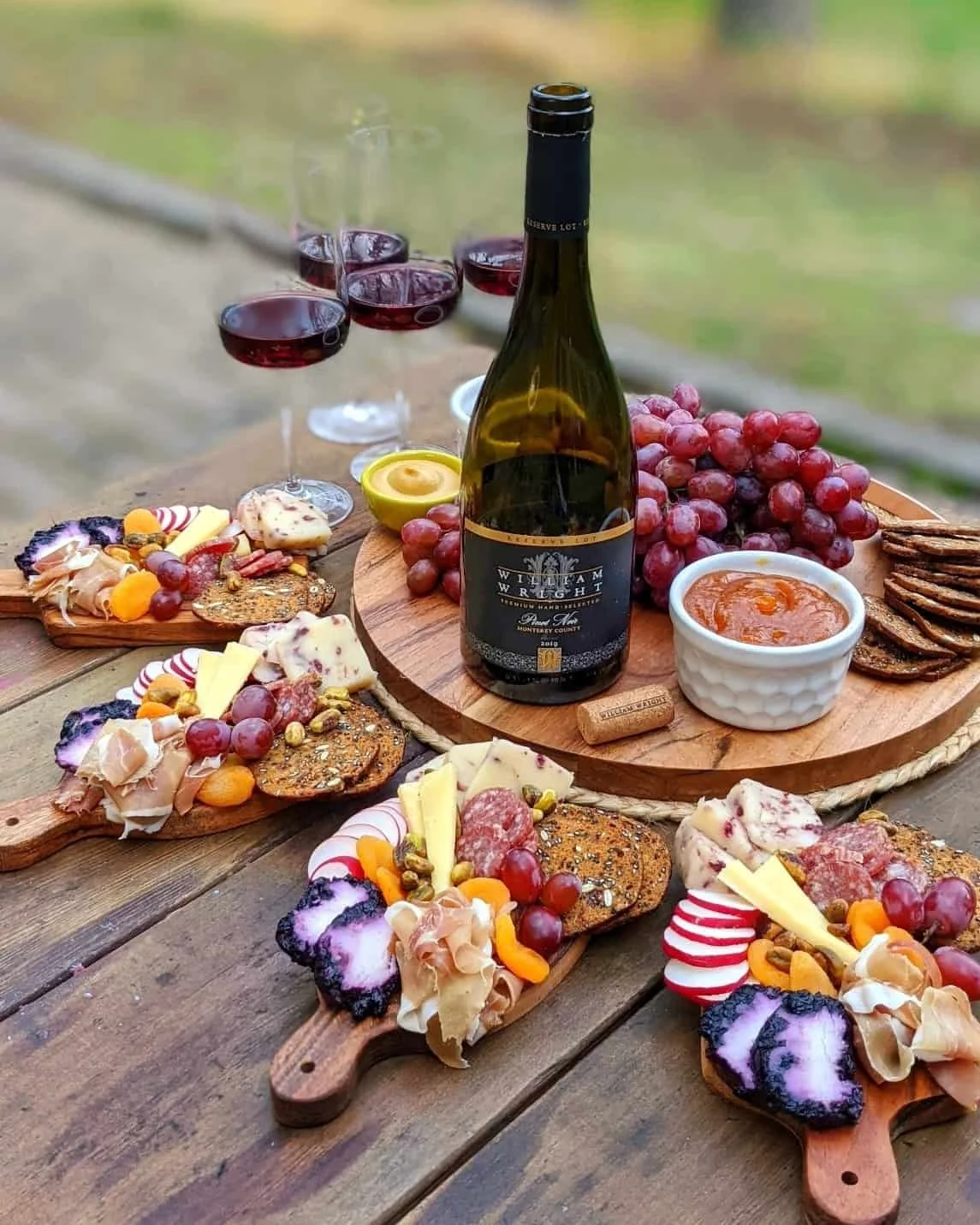 A wine and cheese board spread with grapes, crackers, cheese, and dips, with glasses of red wine on a wooden table outdoors.