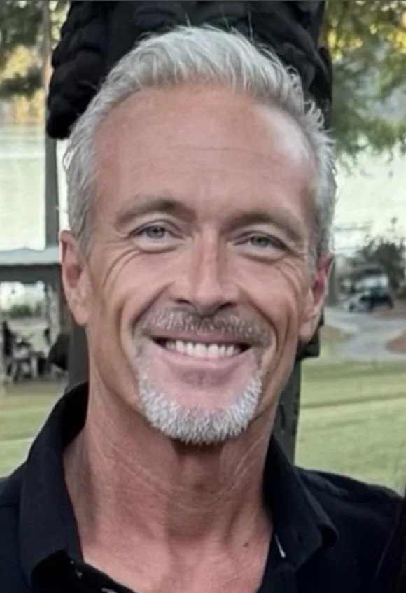 A smiling middle-aged man with short gray hair and a trimmed gray beard, outdoors with trees and a body of water in the background.
