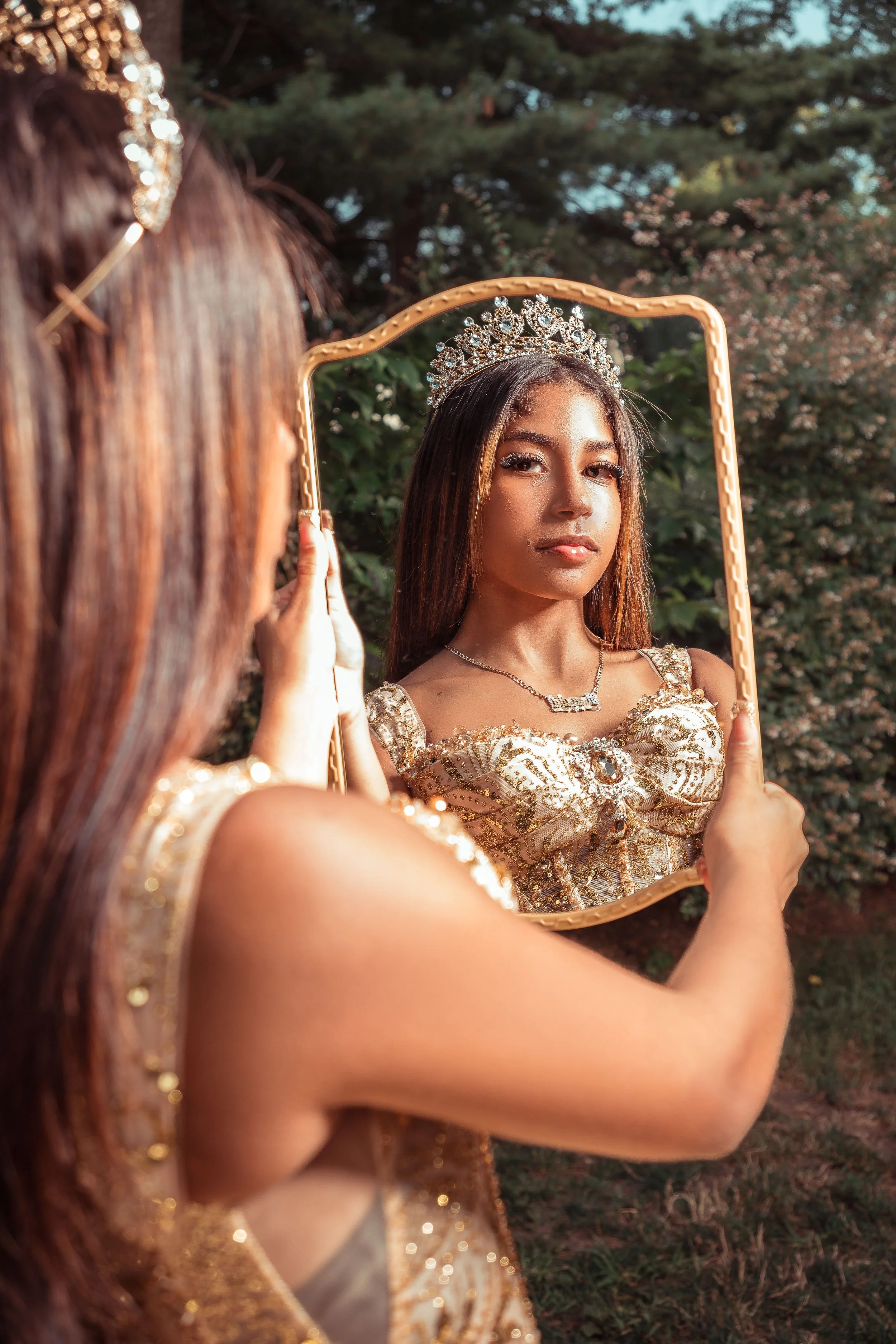 Joven con corona y vestido dorado, mirándose en un espejo en un entorno al aire libre con árboles.