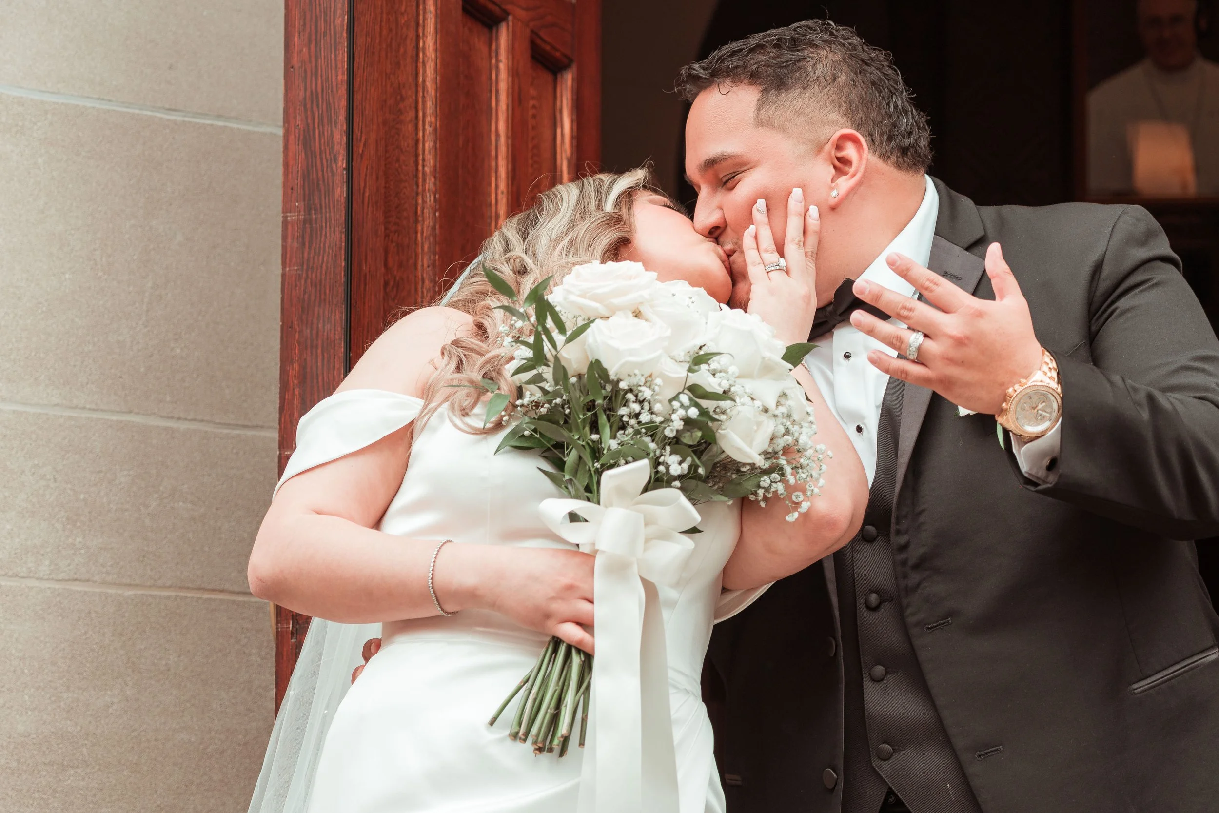 Pareja de novios en su boda, besándose y abrazándose; ella con un ramo de flores blancas; él con traje negro y reloj dorado, con una pared y una puerta de madera al fondo.