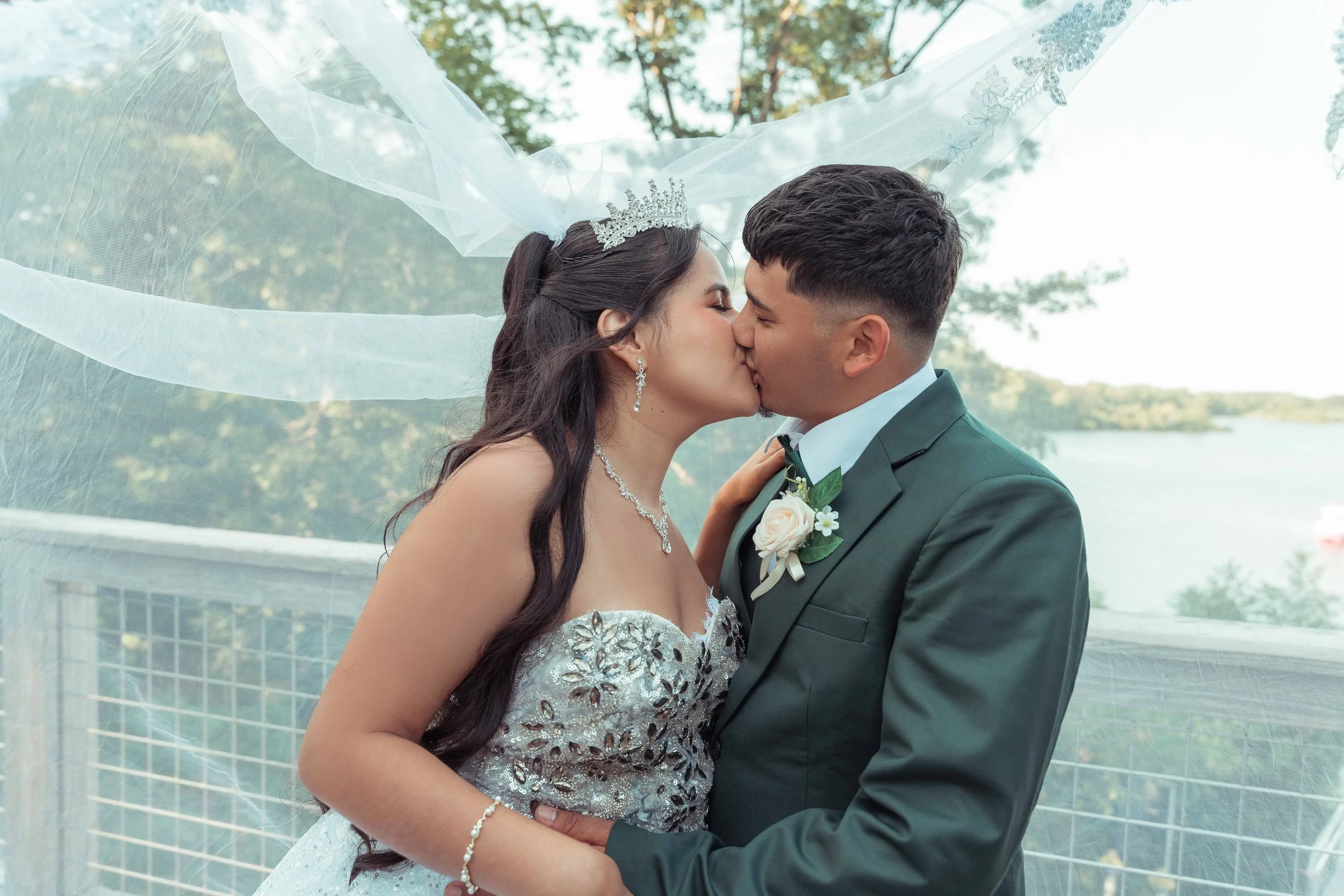 Una pareja de novios en su boda, la novia con tiara y vestido elegante y el novio con traje y flor en el ojal, se besan al aire libre.