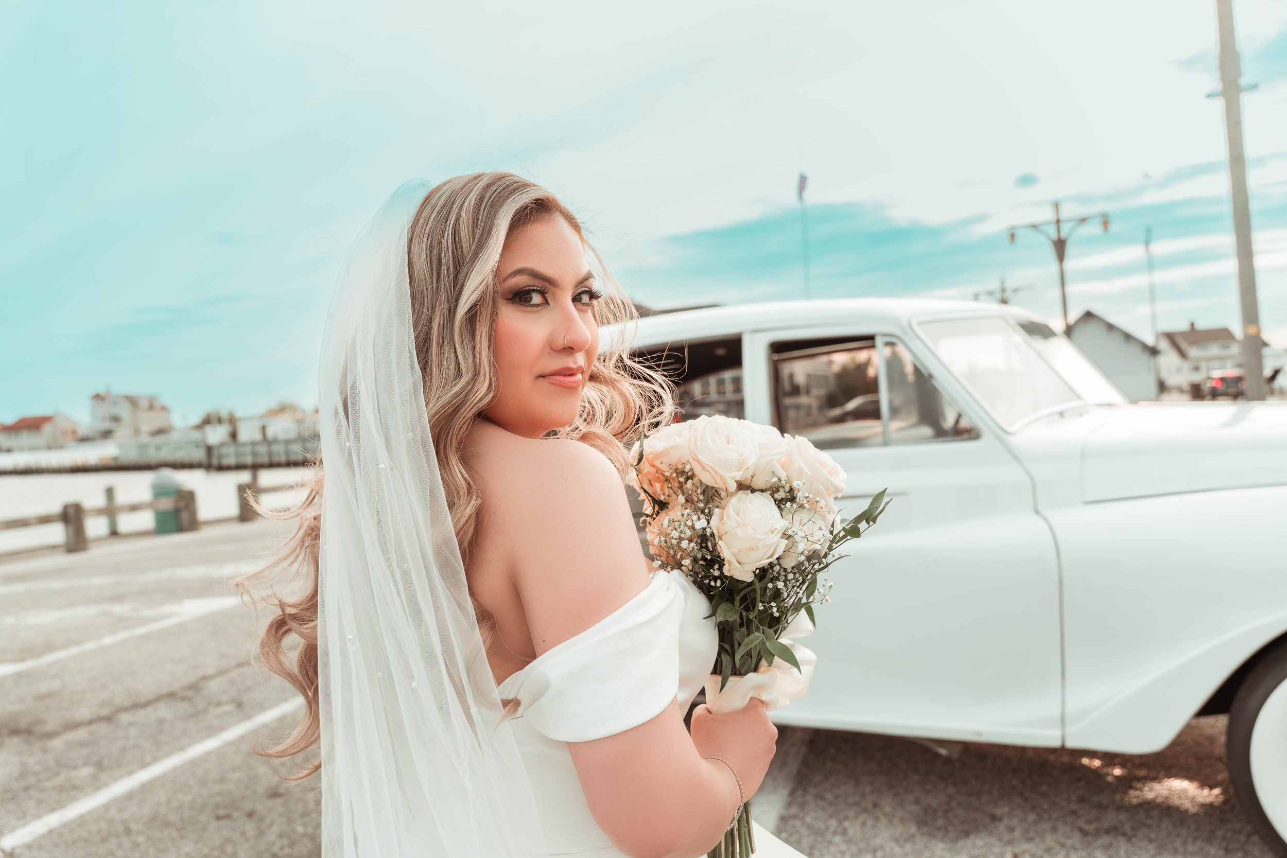 Una mujer con vestido de novia y velo, sosteniendo un ramo de flores, se encuentra junto a un coche clásico en un aparcamiento al aire libre.