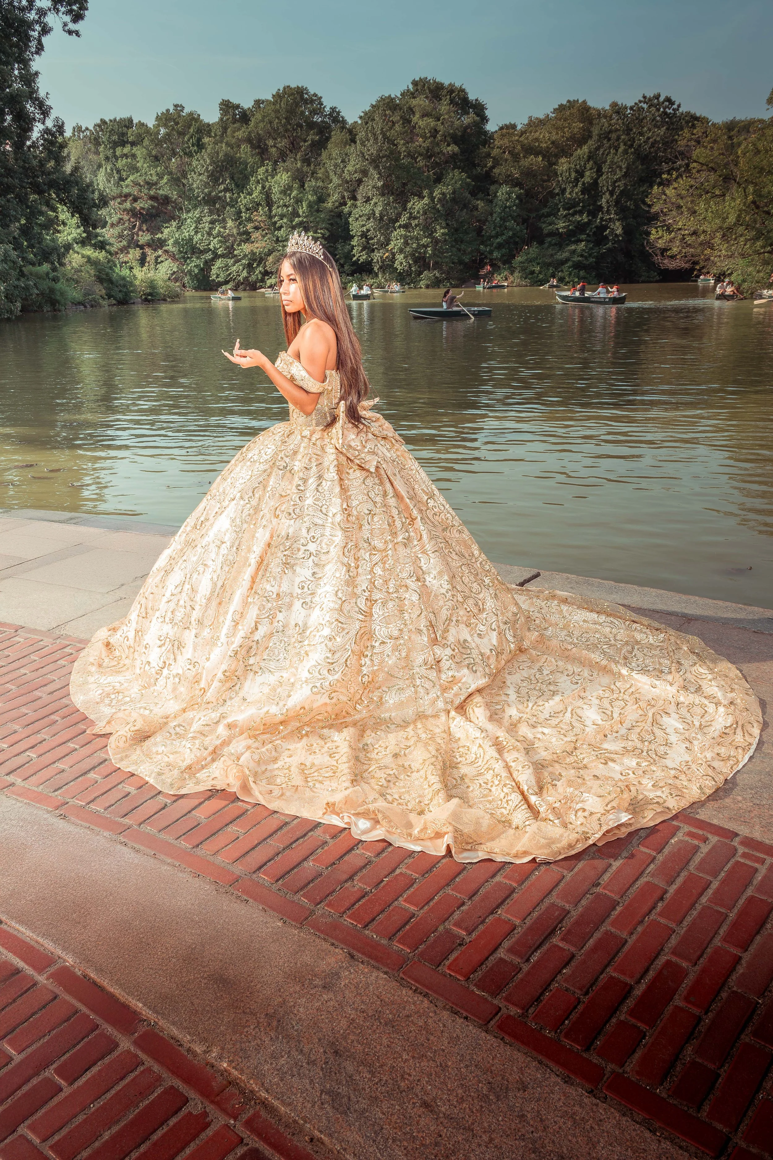 Una joven con vestido de quinceañera y tiara estaba de pie junto a un río, con varias lanchas en el agua y árboles frondosos al fondo.