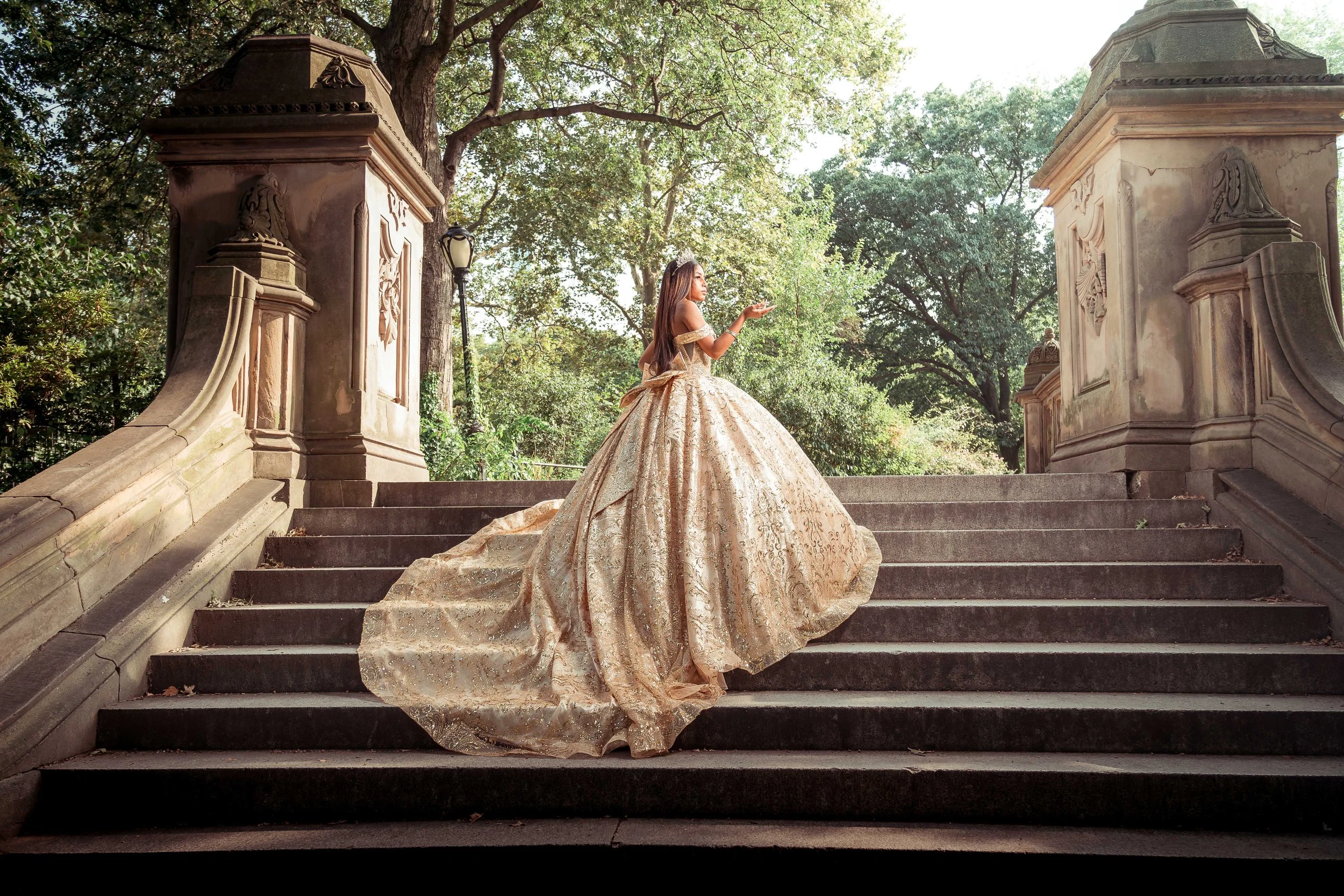 Una mujer con un vestido largo y brillante en las escaleras de piedra de un edificio antiguo, rodeada de árboles y farolas en un parque.