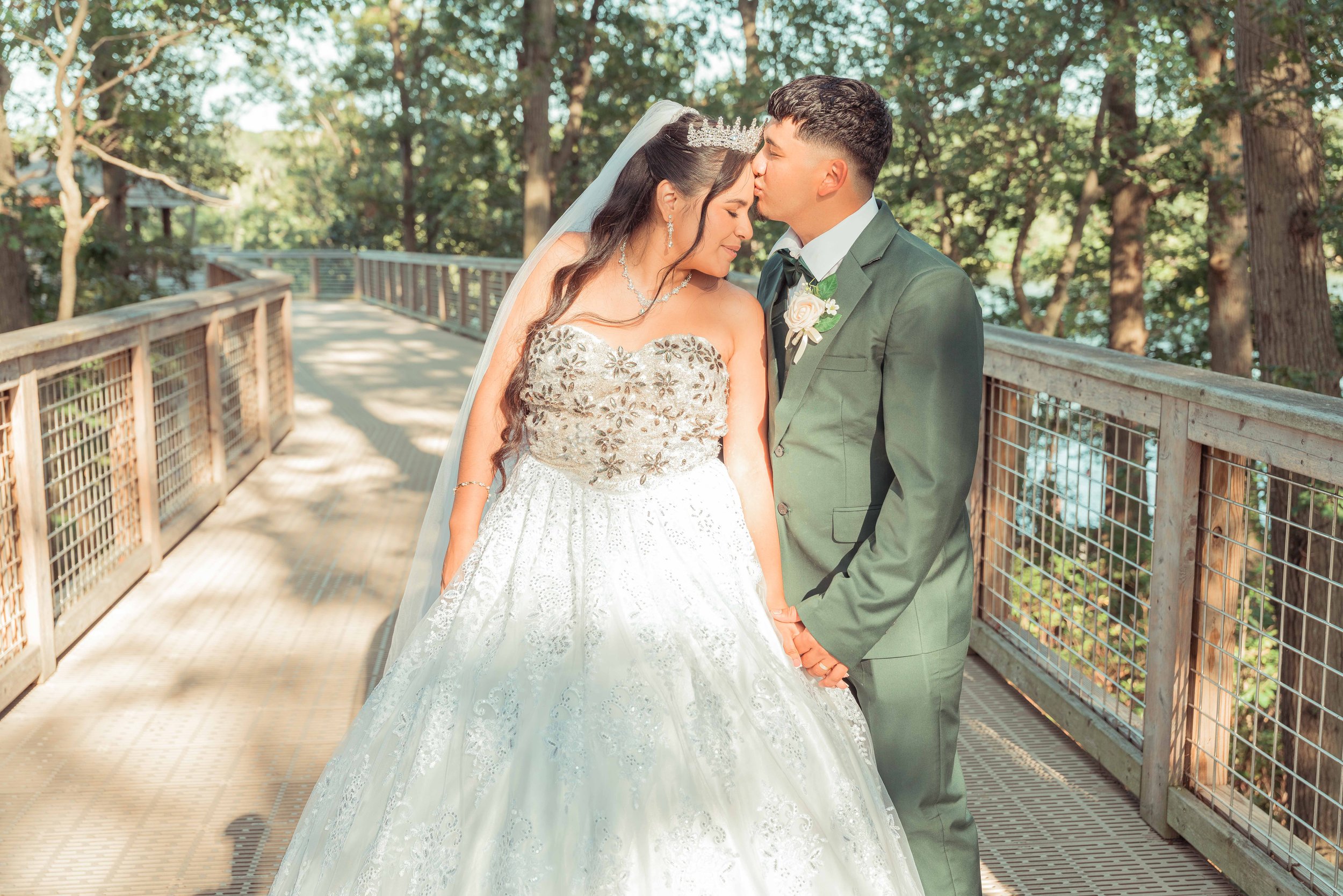 Una pareja de novios en una boda, en la que el hombre besa a la mujer en la frente, en un puente rodeado de árboles.