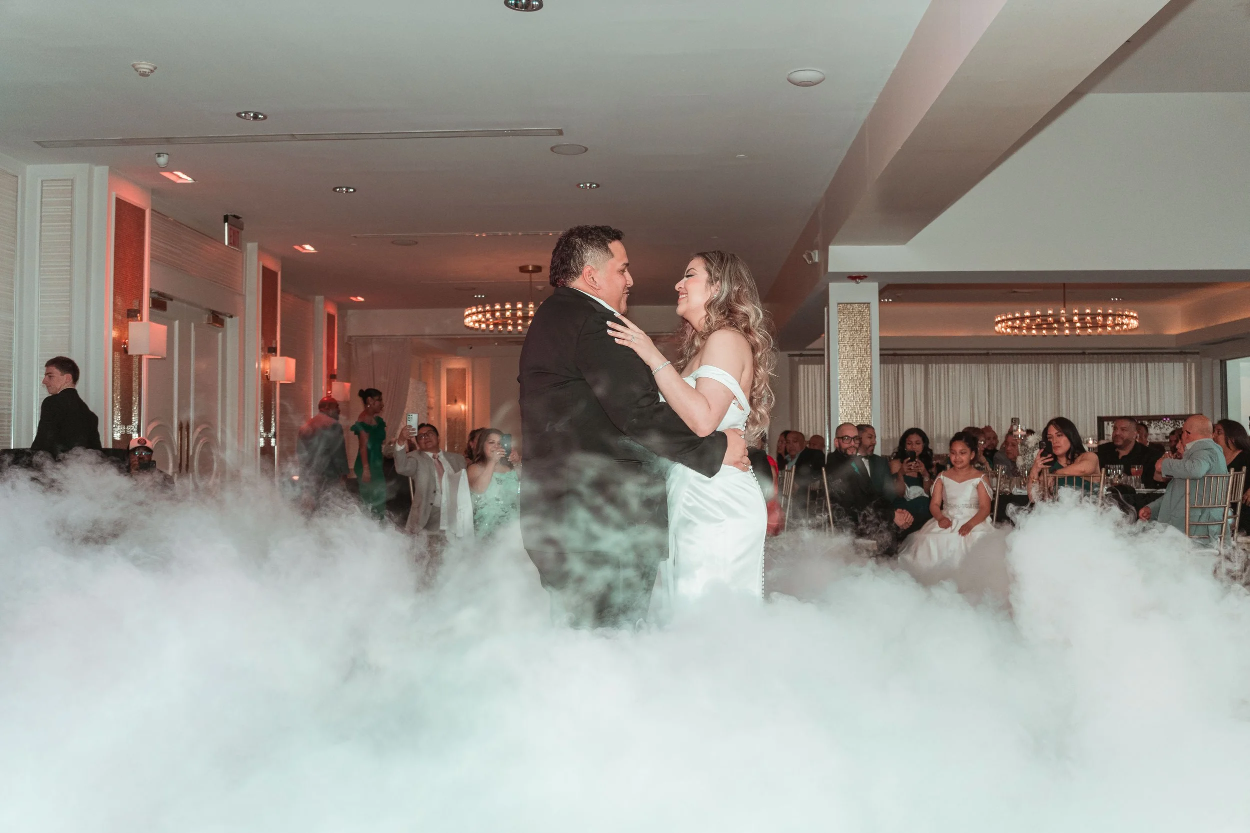 Una pareja bailando en un banquete de boda, rodeada de invitados sentados y tomando fotografías, en un ambiente elegante y romántico.