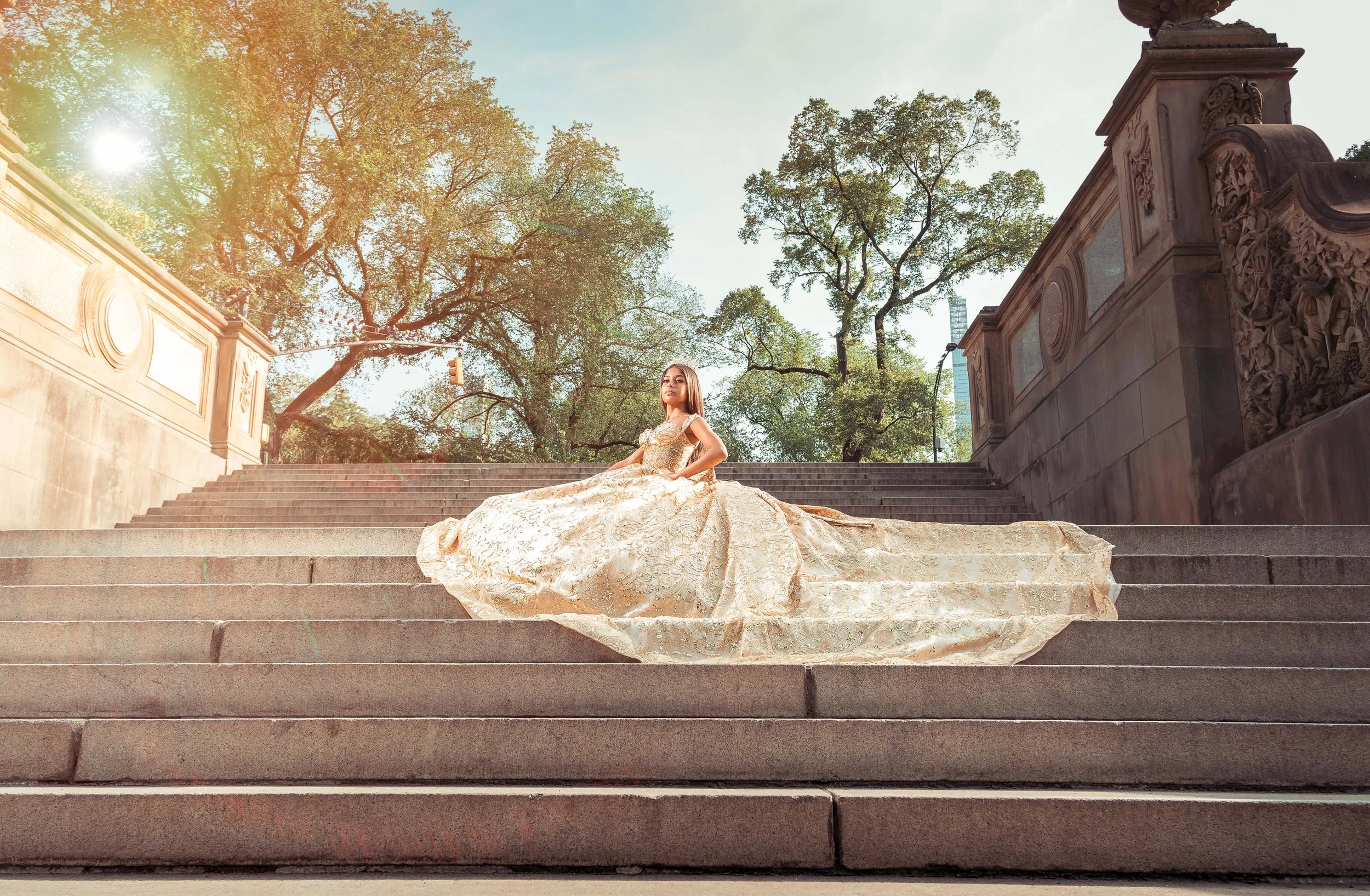 Una mujer con un vestido de novia dorado y largo, sentada en las escaleras de un monumento en un parque, con árboles y un cielo despejado al fondo.
