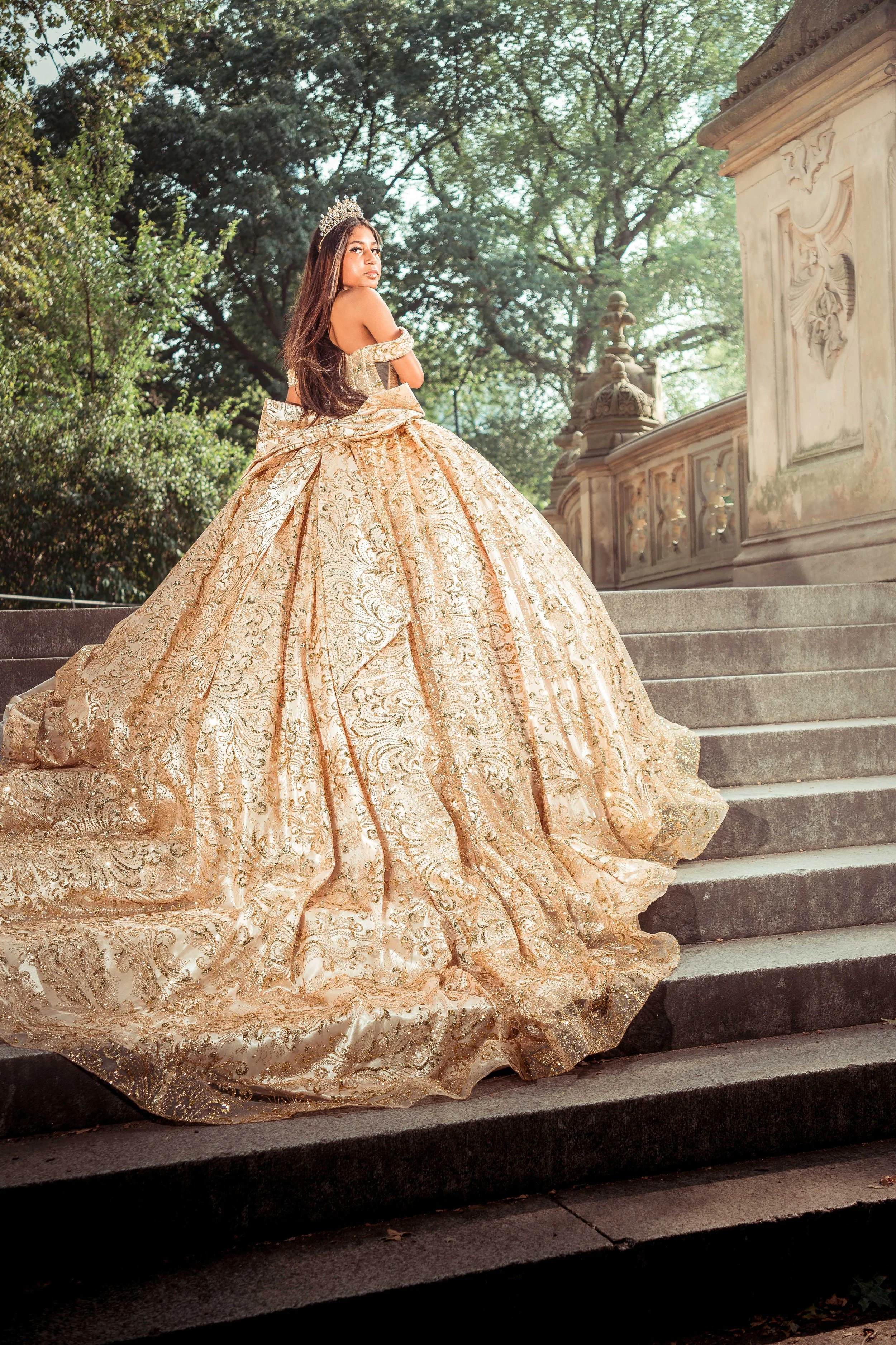 Joven con un vestido de quinceañera dorado con detalles ornamentales y una corona, posando en una escalinata de piedra con árboles y una estructura antigua al fondo.