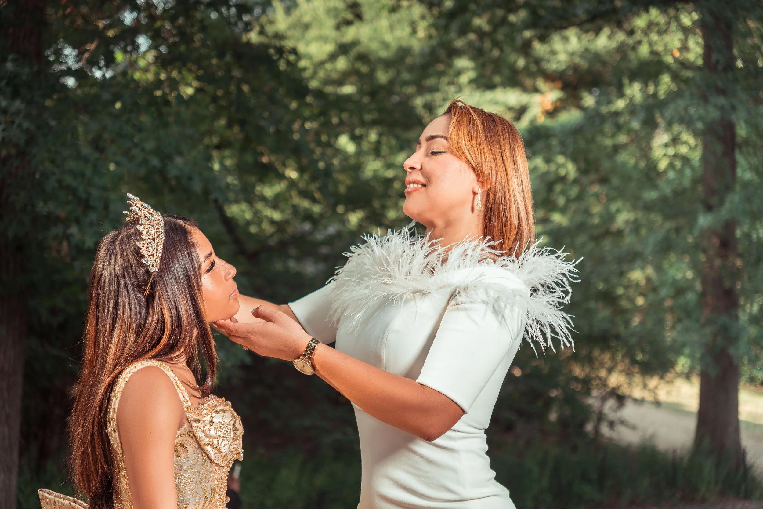 Mujer adulta vestida de blanco, con plumas en los hombros y joyas, que acaricia el cuello de una niña con trenzas, vestida de dorado y con una corona, en un espacio al aire libre con árboles al fondo.