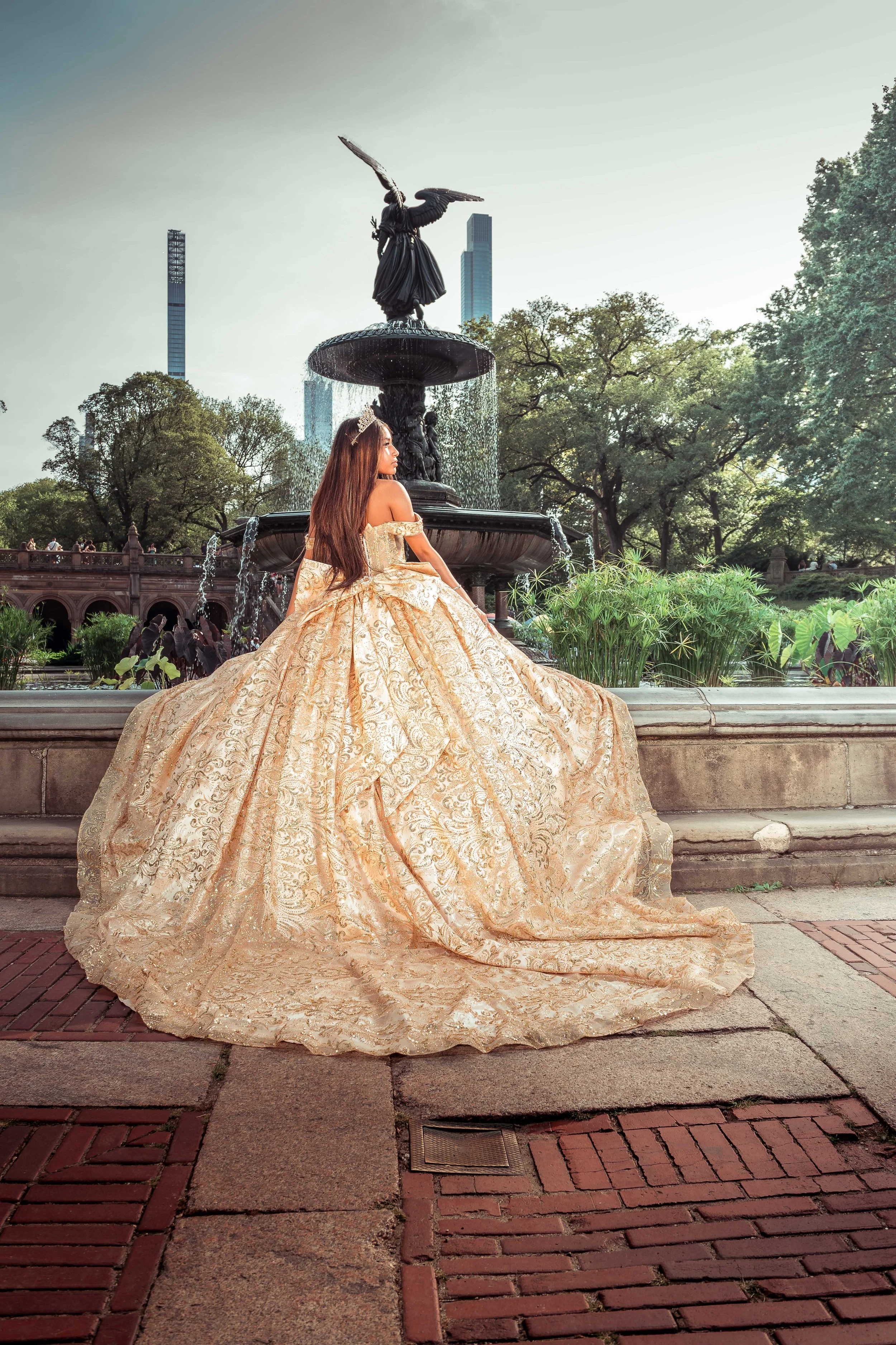 Una joven con un vestido de novia dorado y una diadema en el pelo, sentada frente a una fuente en un parque, con edificios altos al fondo.