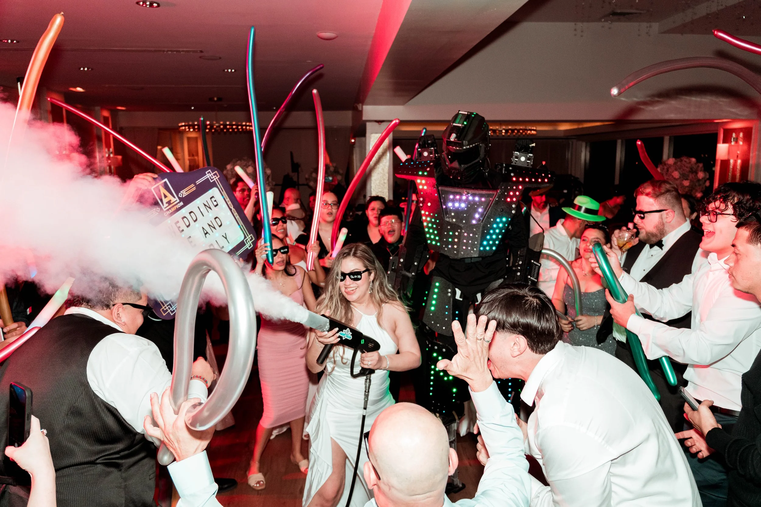 Personajes disfrazados de robots luminosos y gente bailando en una fiesta con un ambiente festivo, globos y confeti.