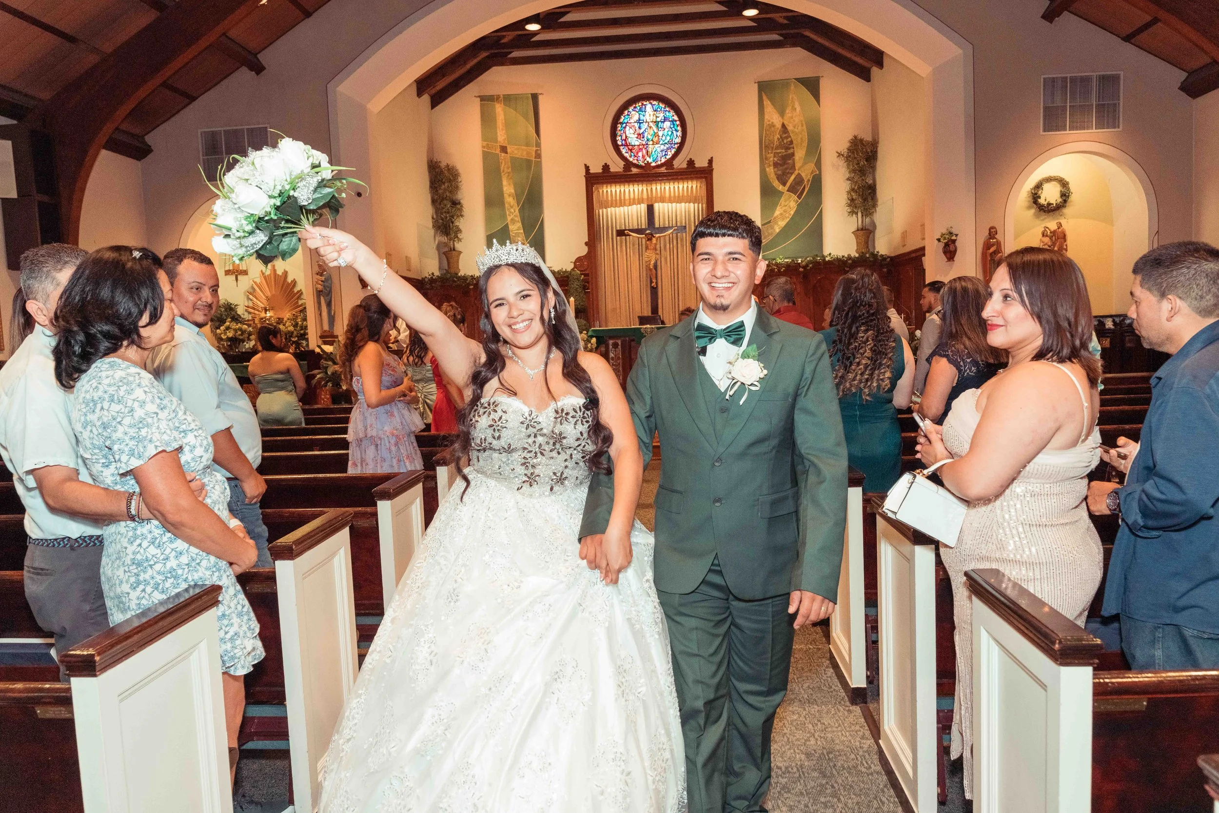 Una pareja de recién casados en la iglesia; la novia lleva un vestido de novia y una diadema con velo, y el novio lleva un traje verde con pajarita, rodeados de familiares y amigos durante la ceremonia.