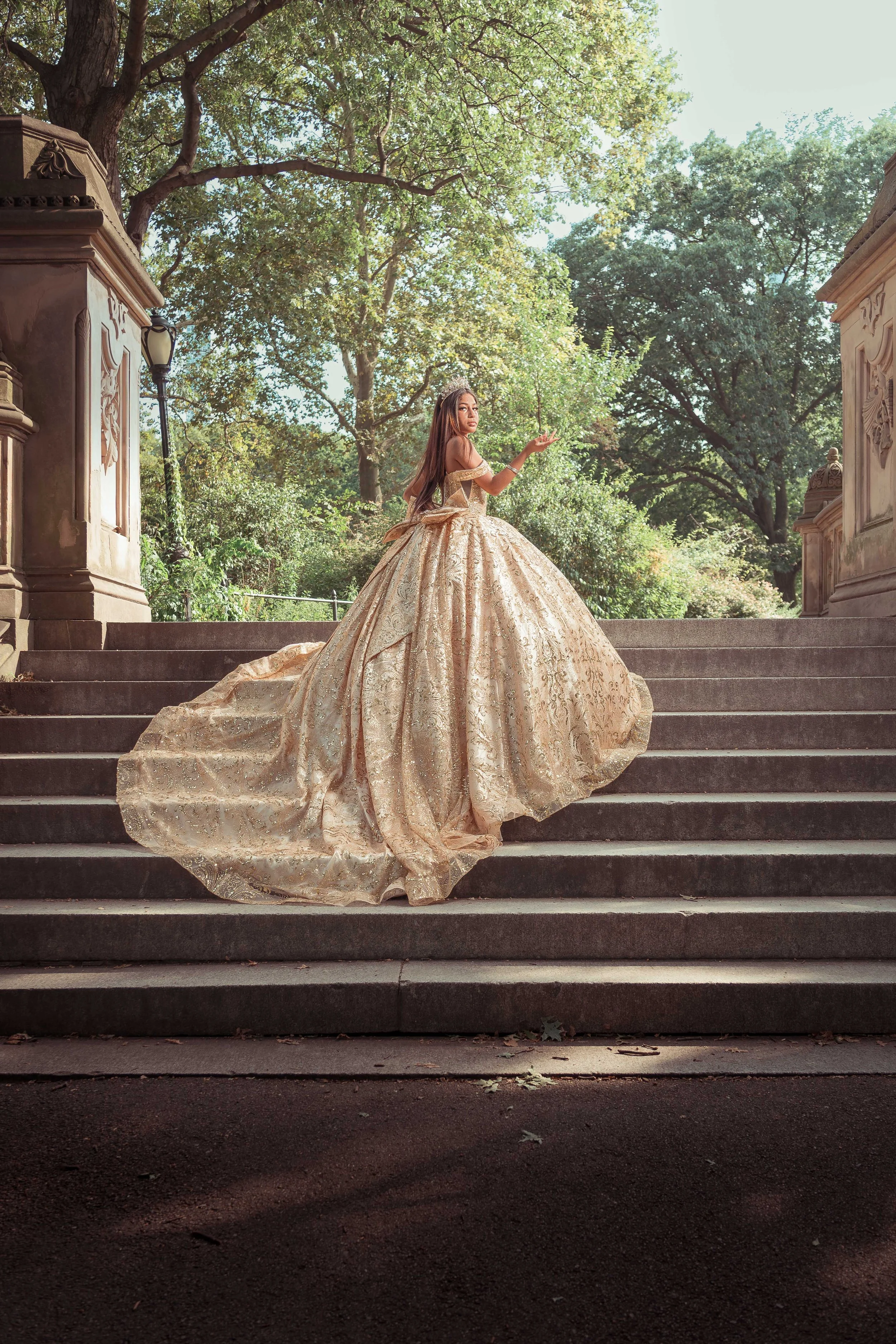 Una mujer vestida con un elegante vestido dorado de corte amplio, de pie en medio de unas escaleras de piedra, con árboles verdes al fondo y la luz natural del día.