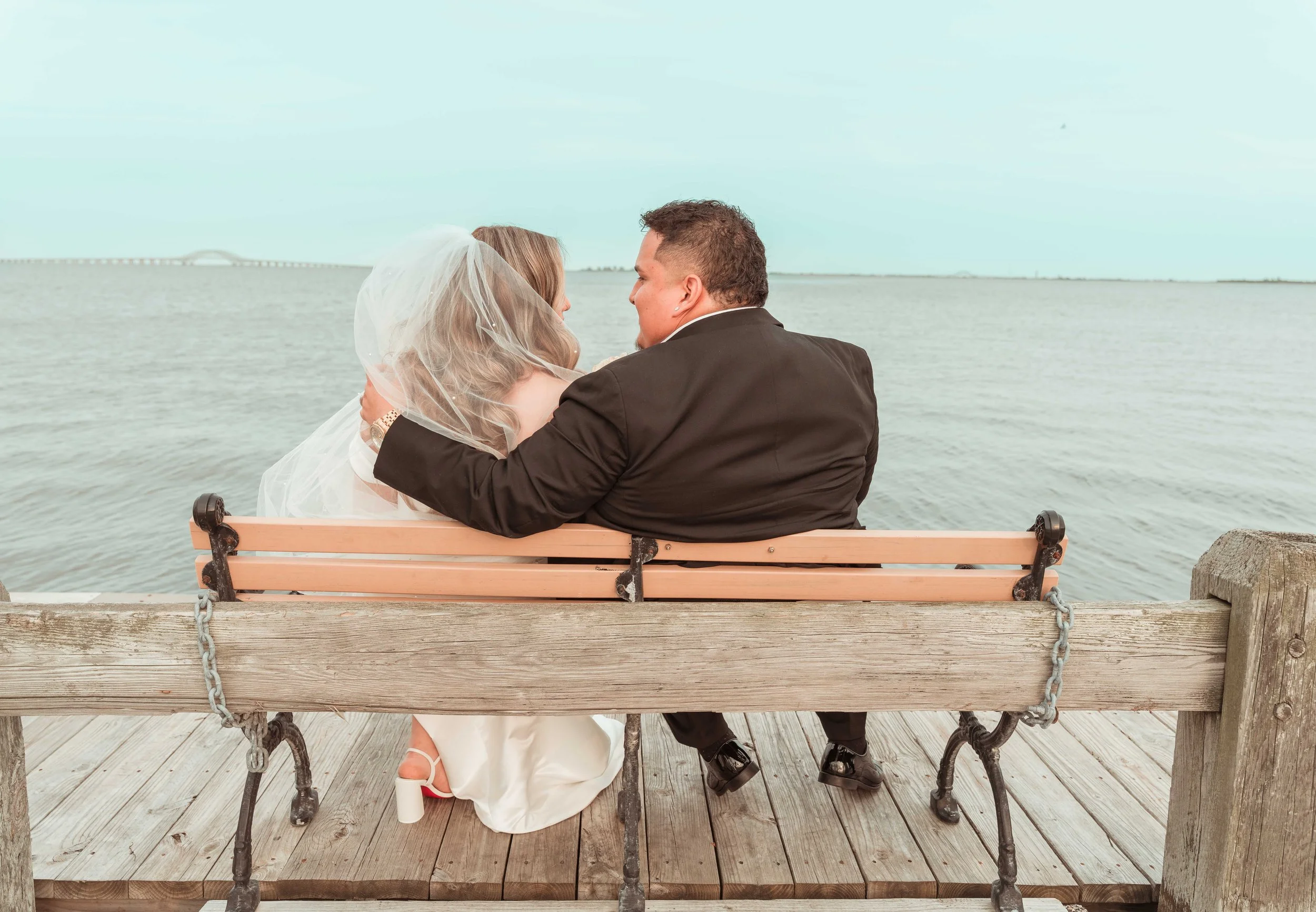 Pareja vestida de boda sentada en un muelle junto al agua, con un puente visible al fondo, en un día despejado.