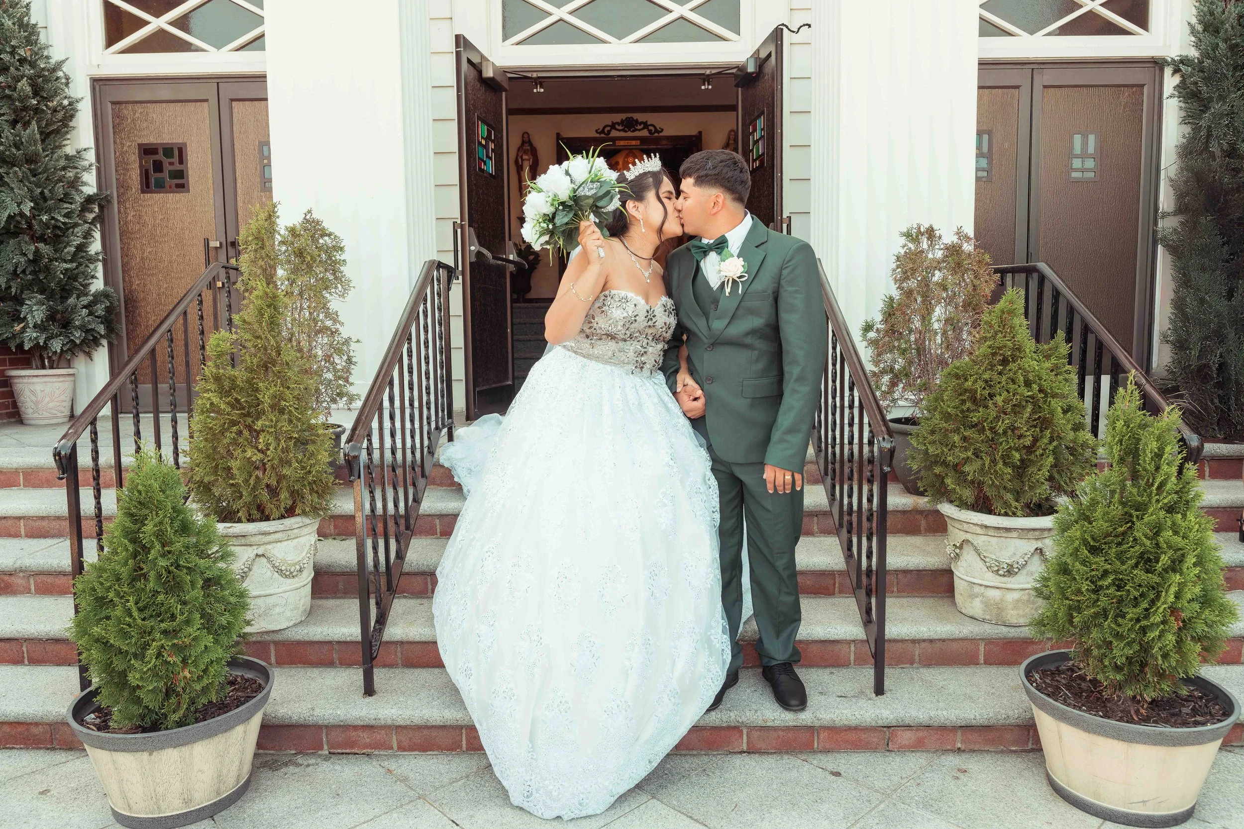 Una pareja de novios besándose a la entrada de una iglesia; la novia lleva un vestido de novia y una corona, y el novio, un traje y una pajarita; ambos están rodeados de macetas y plantas.