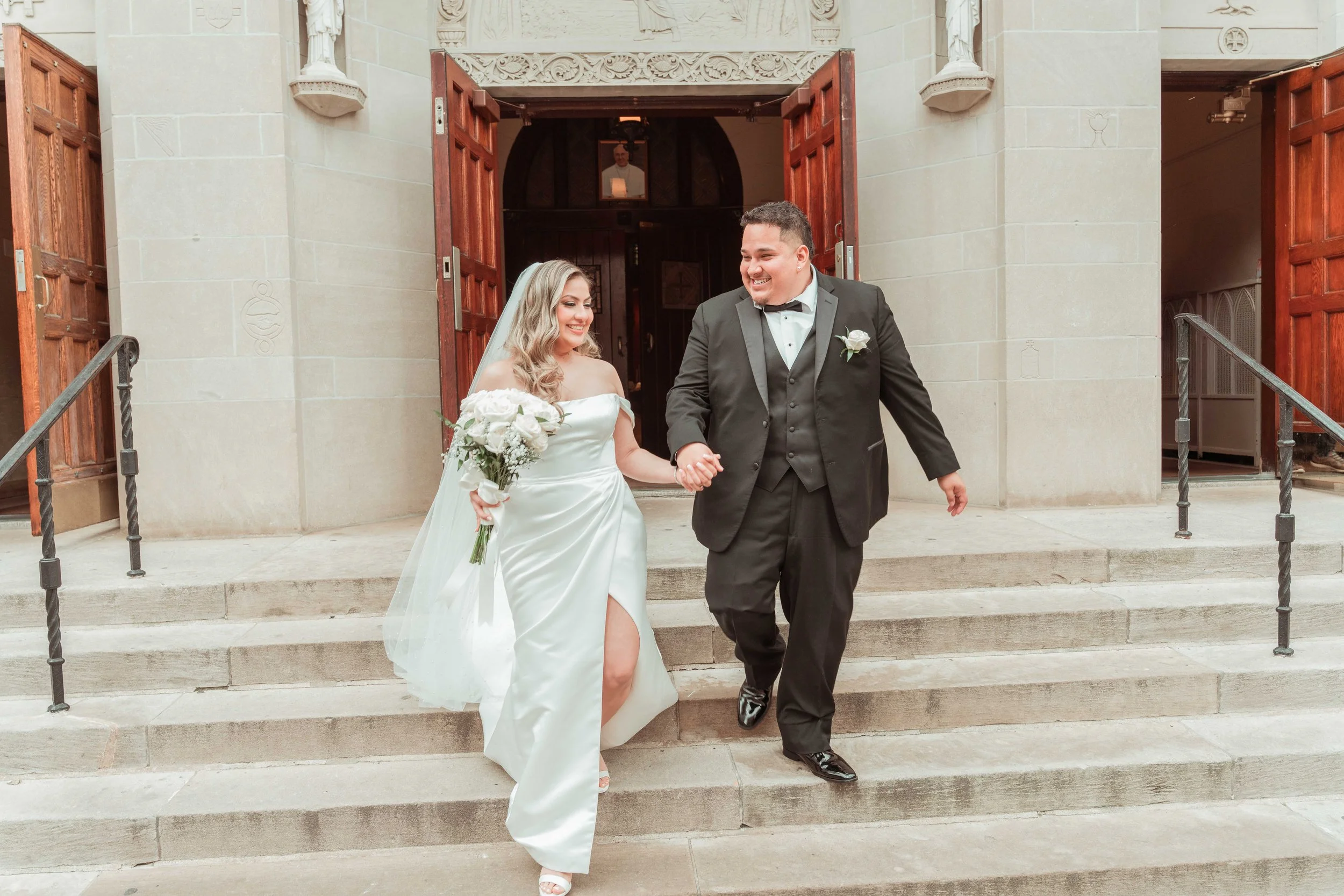 Se ve a una pareja de novios saliendo de una iglesia católica. La novia lleva un vestido de novia blanco y sostiene un ramo de flores blancas, y el novio lleva un traje negro con pajarita y una flor en la solapa. Ambos sonríen y van de la mano, b