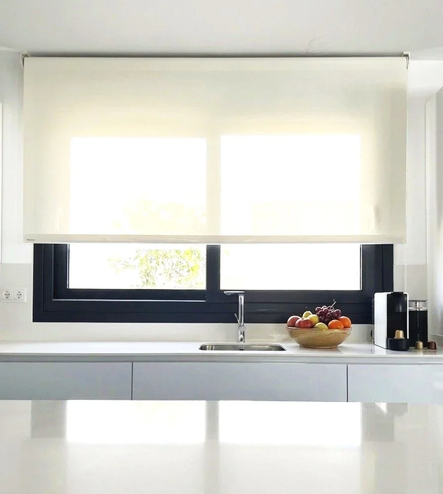 Ventana grande con cortina ligera sobre fregadero y encimera de cocina con fruta y una máquina de café.