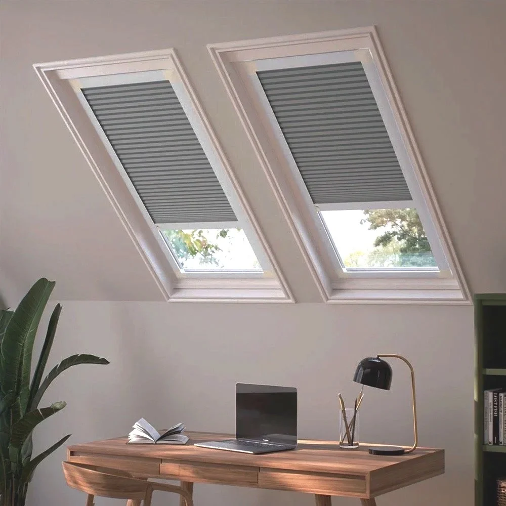 Estudio con dos ventanas grandes en el techo, persianas bajadas parcialmente, escritorio de madera con una laptop, libro, lámpara y utensilios de escritura, y planta a la izquierda.