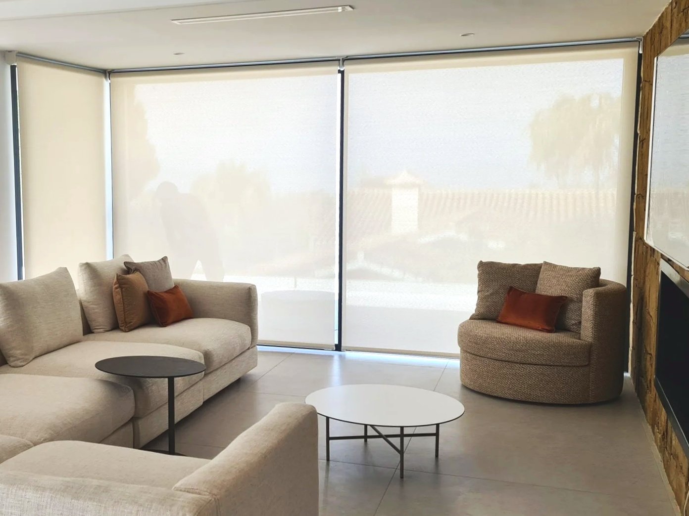 Sala con sillón beige, sillón redondo con cojines, mesa negra, mesas blancas, cortinas blancas translucidas y muro de madera.