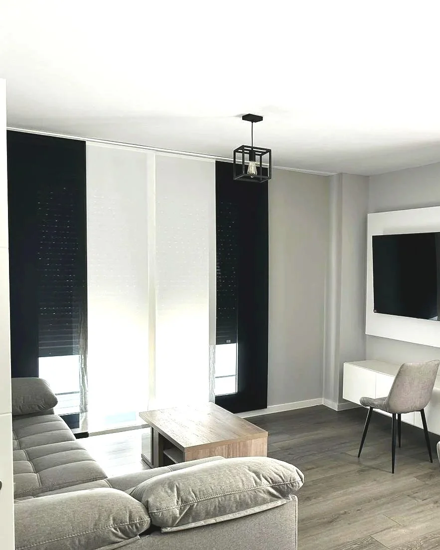 Sala de estar moderna con sillón gris, mesa de centro de madera, cortinas blancas y negras, televisor en la pared y lámpara colgante negra.
