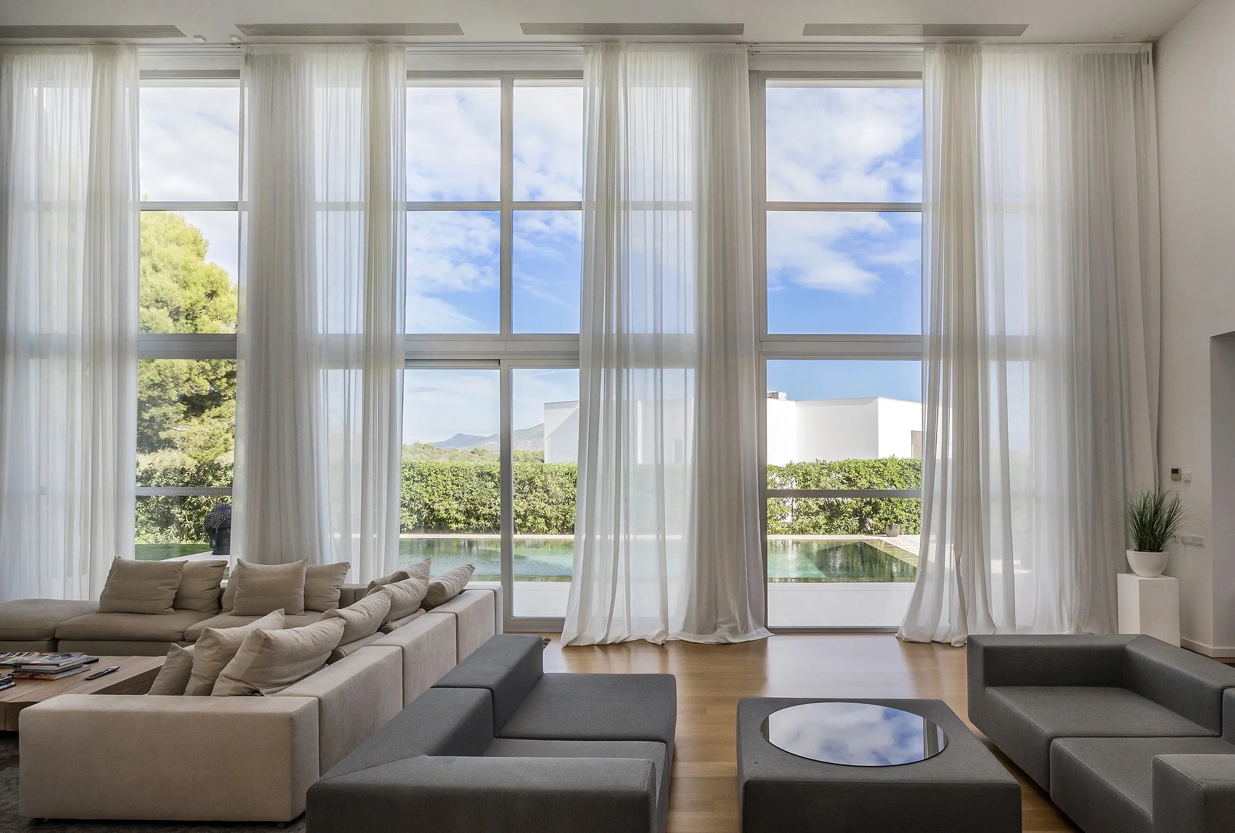Sala moderna con ventanas grandes y cortinas blancas, vista a jardines y montañas, muebles en tonos neutros.
