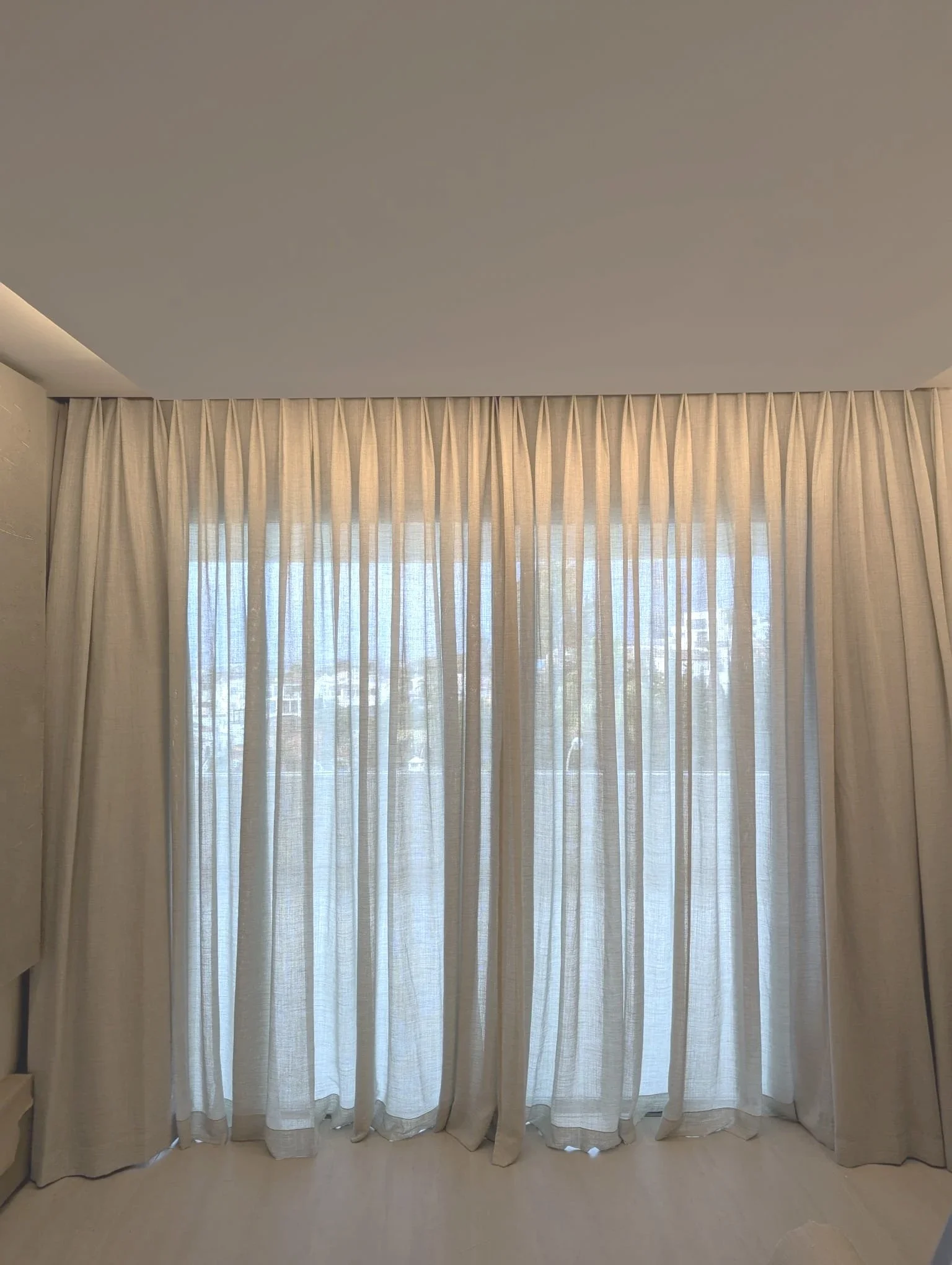 Cortinas blancas cubren una ventana grande con vista a edificios y cielo.