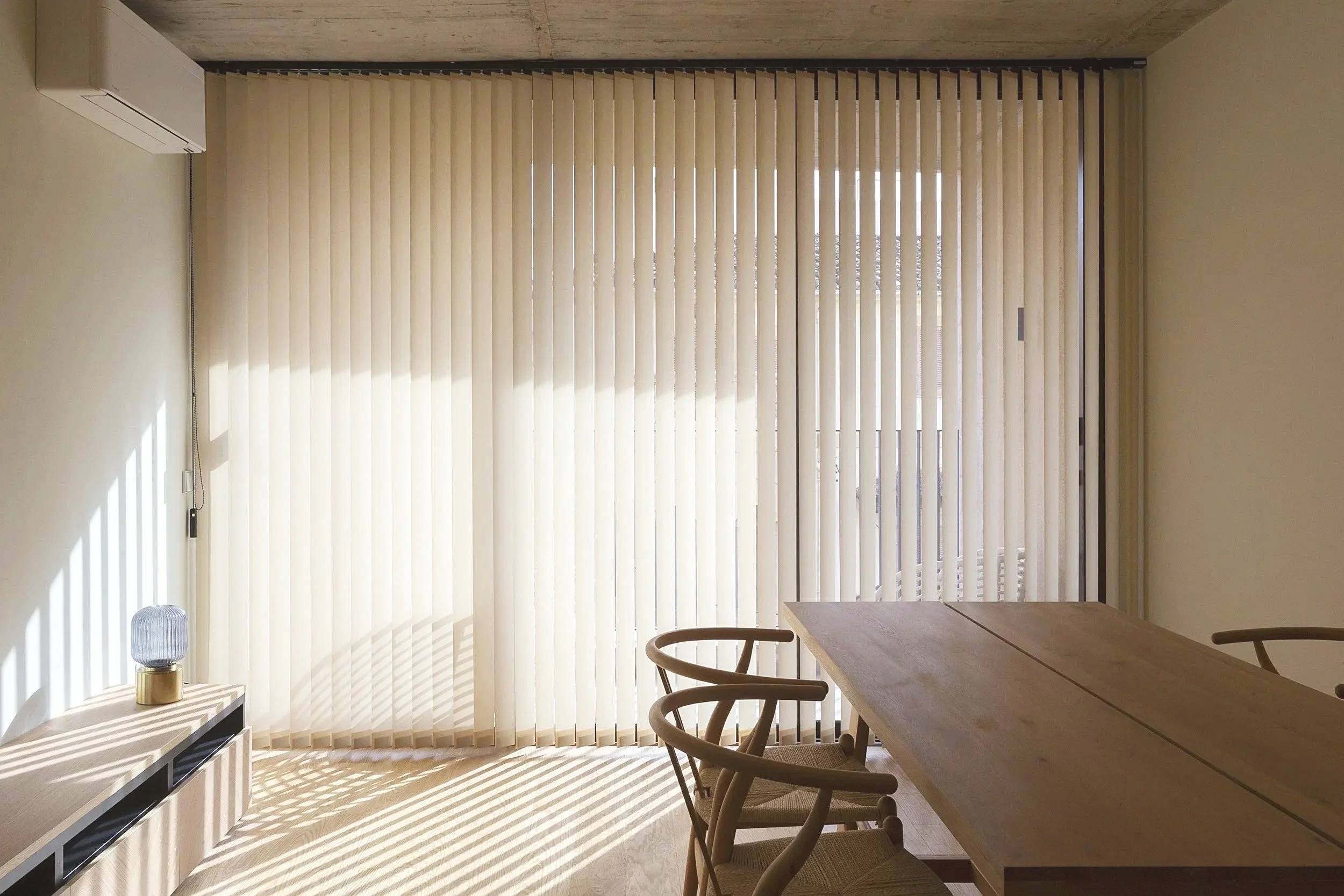 Interior de una sala con cortinas de persiana beige, mesa de madera, sillas, una lámpara y una unidad de aire acondicionado en la pared, con luz del sol proyectada a través de la persiana.