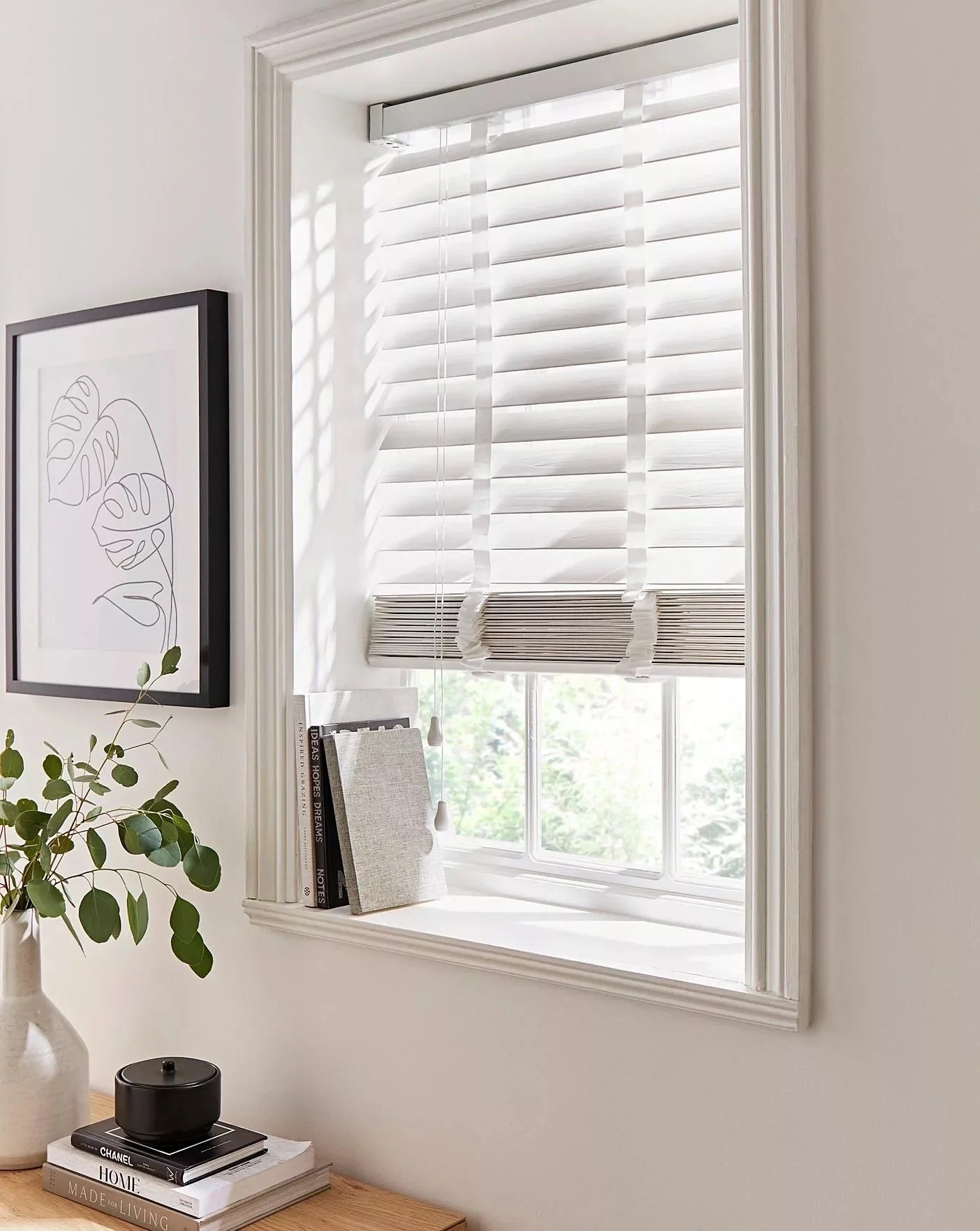 Interior de una habitación con una ventana de persianas blancas, un perchero con libros y una silla, y una planta en una mesa