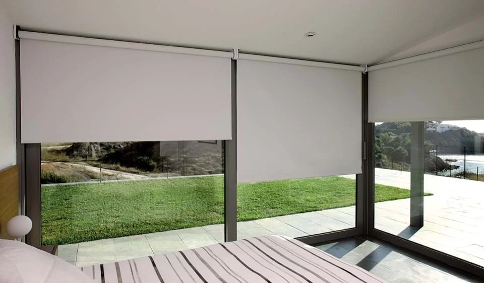 Ventanas grandes con persianas enrollables blancas en habitación con vista a un jardín y vista al mar en el fondo.