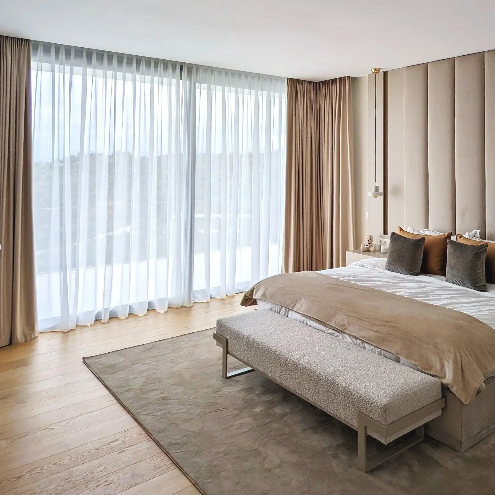 Una habitación de hotel con paredes beige, cortinas blancas y beige, un ventanal grande con vista a la naturaleza, una cama con sábanas blancas y almohadas beige y gris, un banco al pie de la cama y una alfombra gris