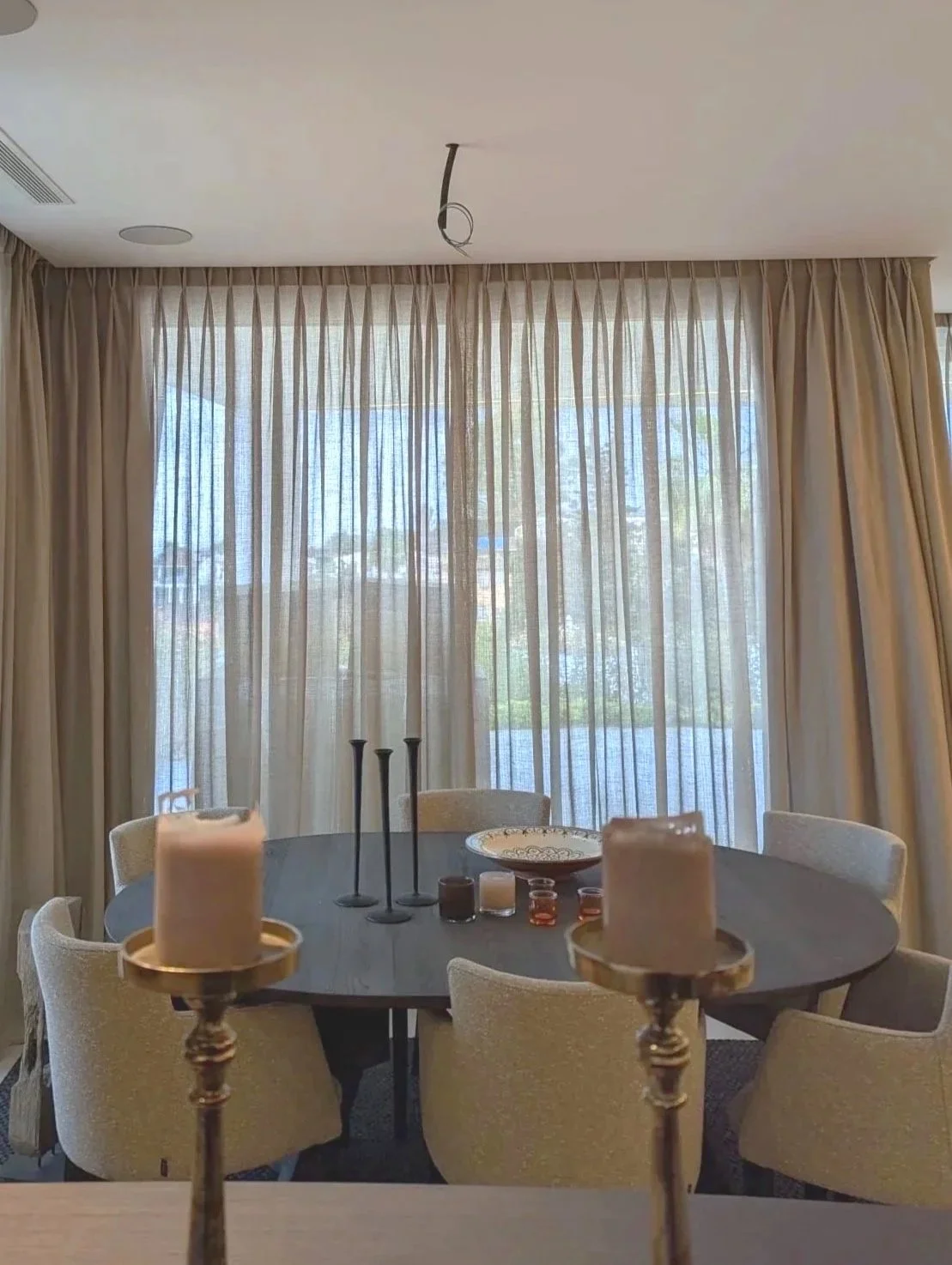 Vista de una sala de comedor con cortinas largas y translucidas, mesa redonda con sillas beige, y decoración con velas en el centro.