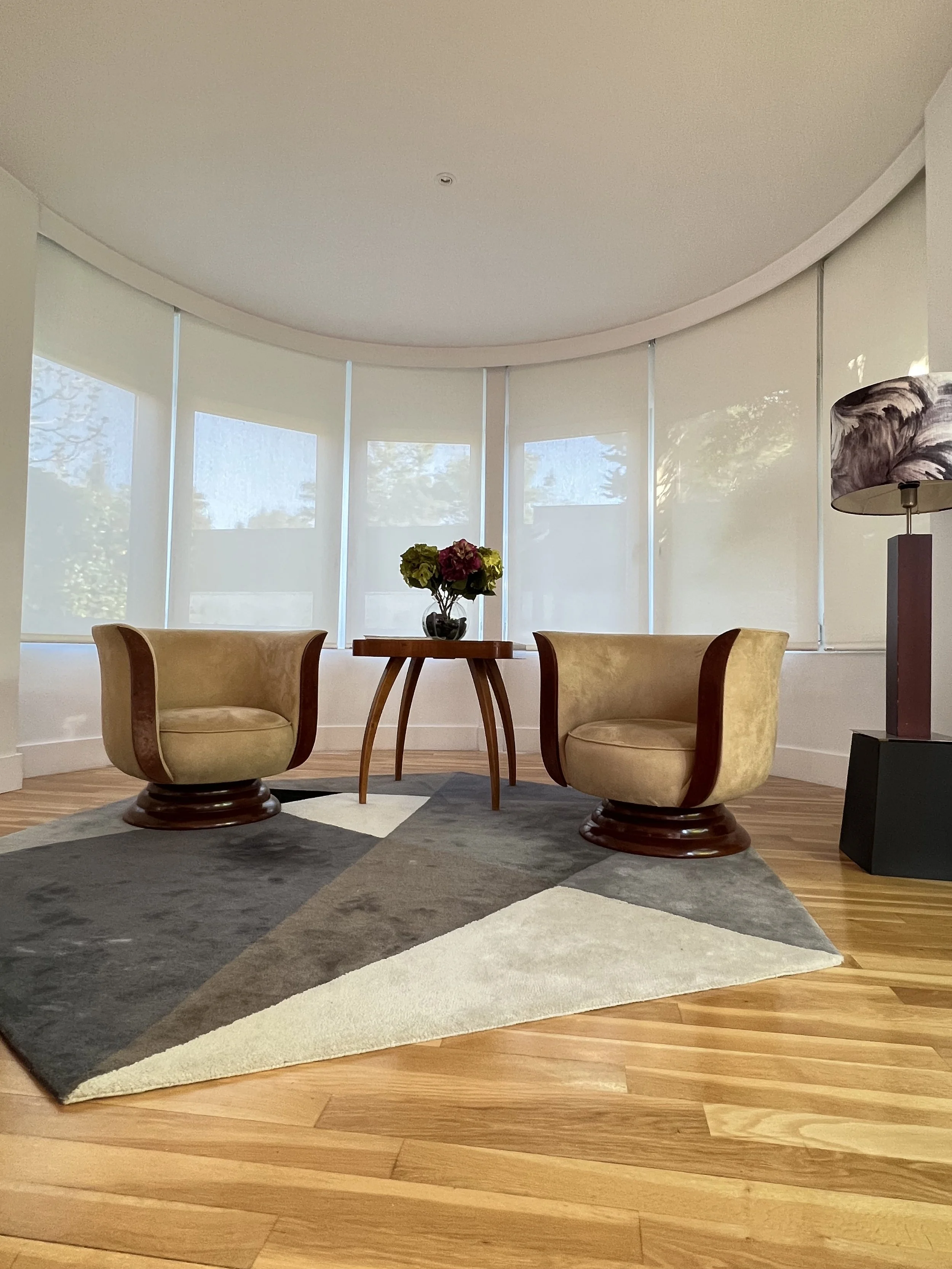 Sala de estar con dos sillones beige, una mesa de madera con un florero con flores y una lámpara de pie con pantalla de diseño abstracto, alfombra en patrón geométrico de colores neutrales y madera clara en el suelo, con ventanas con cortinas blancas y vistas de árboles exteriores.