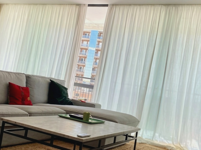Sala de estar con sofá gris, cojines rojos y verdes, mesa de centro de mármol, cortinas blancas y vista de un edificio alto.