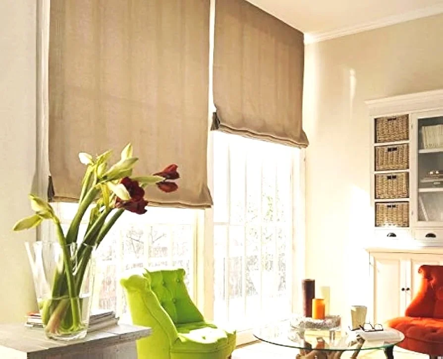 Sala de estar con ventanas grandes con cortinas beige, sillones verdes y naranjas, mesa de centro de cristal, y una repisa blanca con cestas de mimbre.