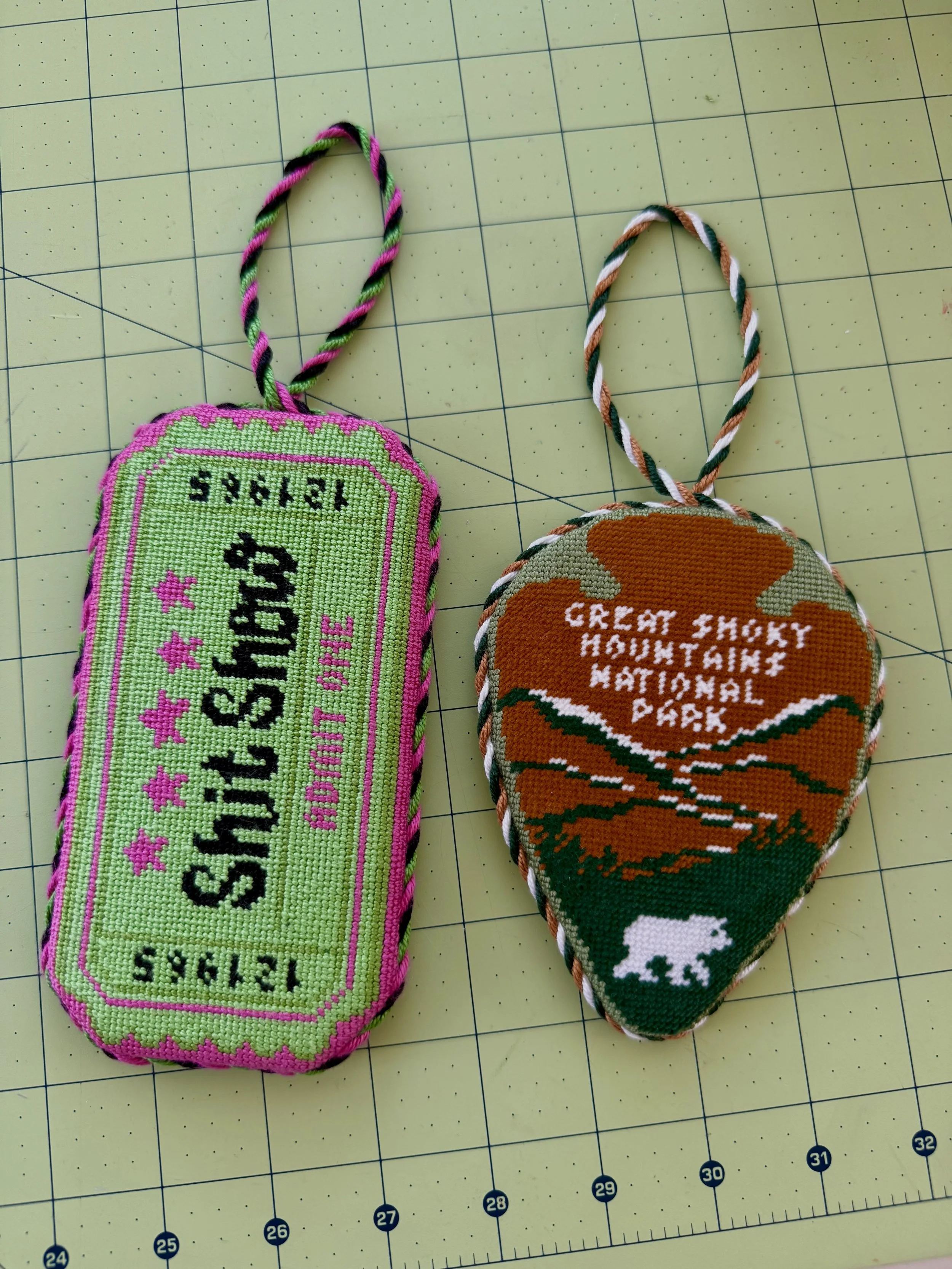 Two needlepoint keepsakes, one shaped like a show ticket, one like a National Park sign.