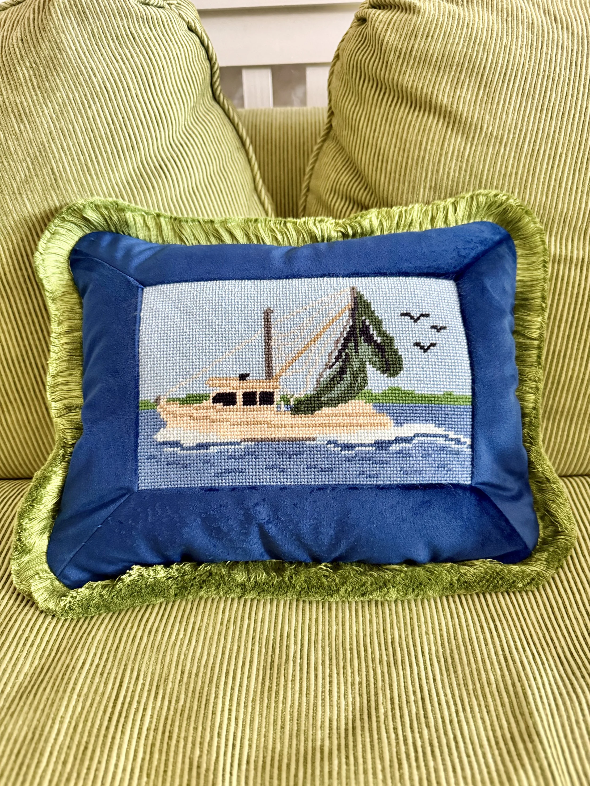 A blue cushion with a needlepoint picture of a boat, a pine tree, and birds, placed on a green velvet armchair.